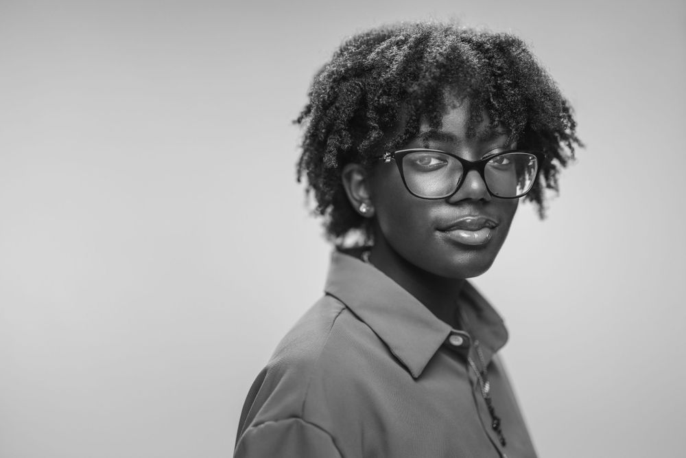 A monochrome studio portrait of someone wearing glasses with natural curly hair and a collared shirt.