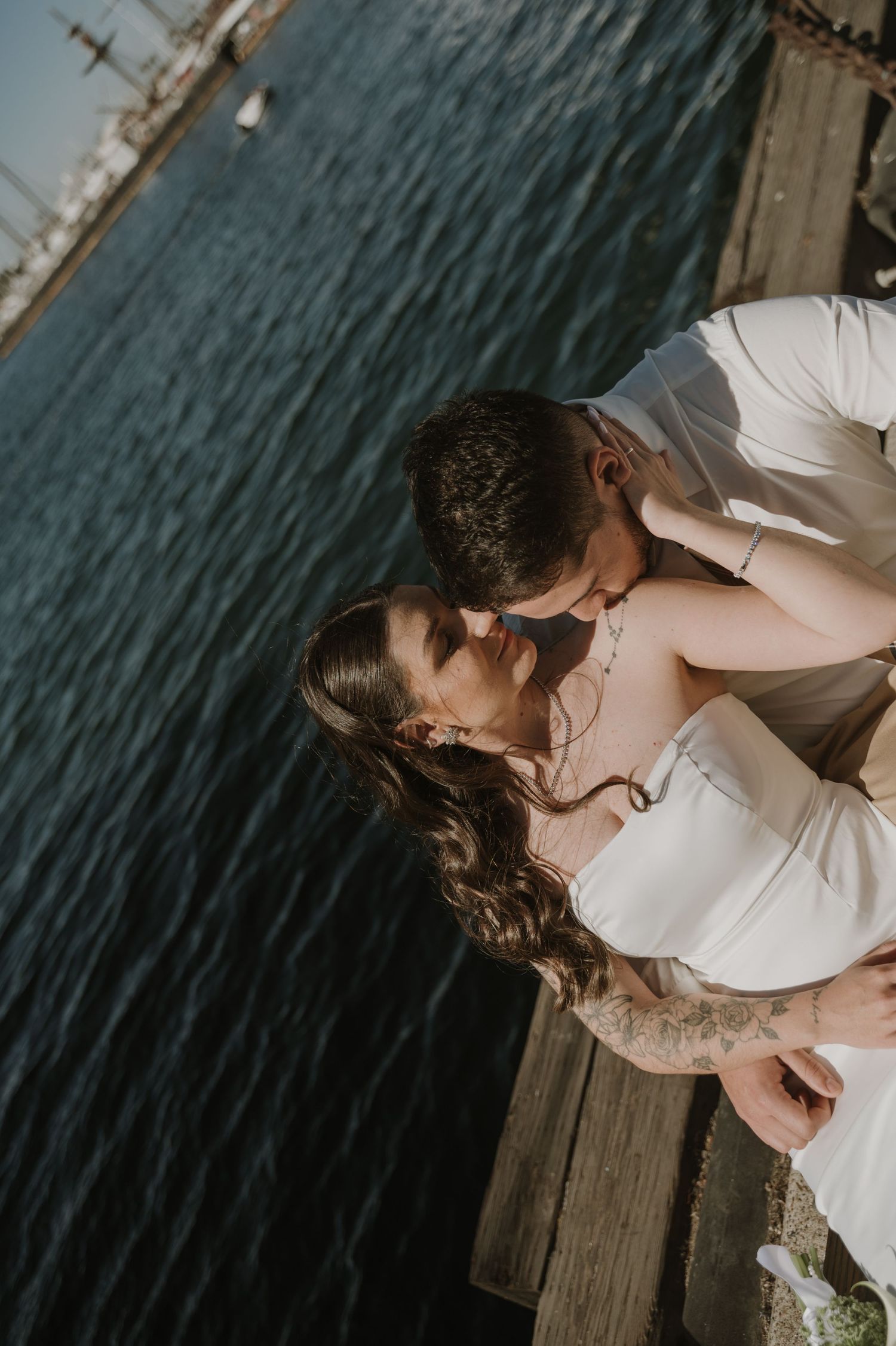 A romantic couple in white embraces on a wooden dock by the water.