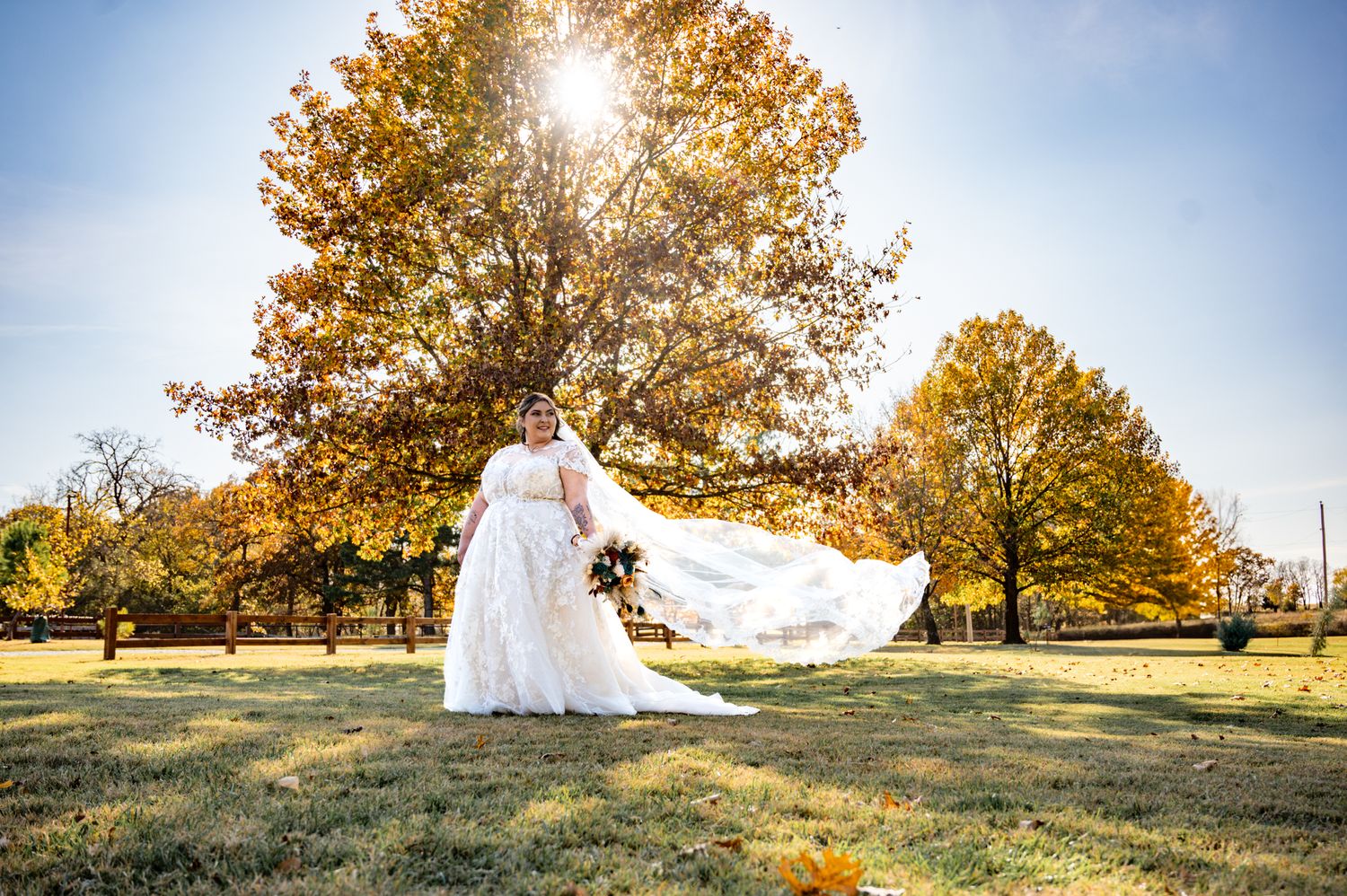 A bride's veil flows dramatically in front of golden autumn trees on a sunny day.