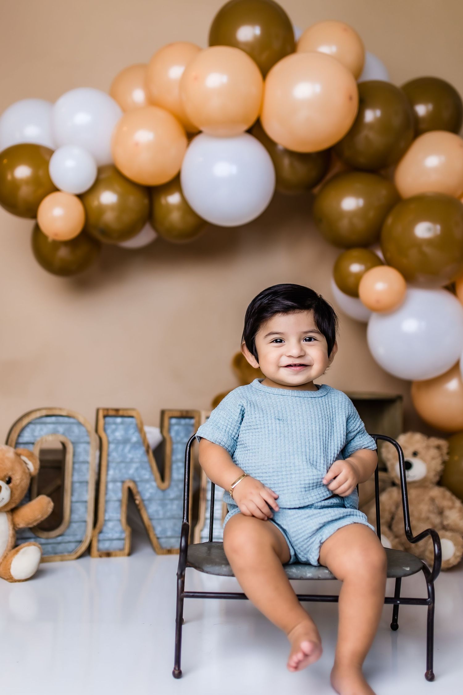 A baby in blue romper sits on chair under balloon arch with ONE letters for first birthday celebration.