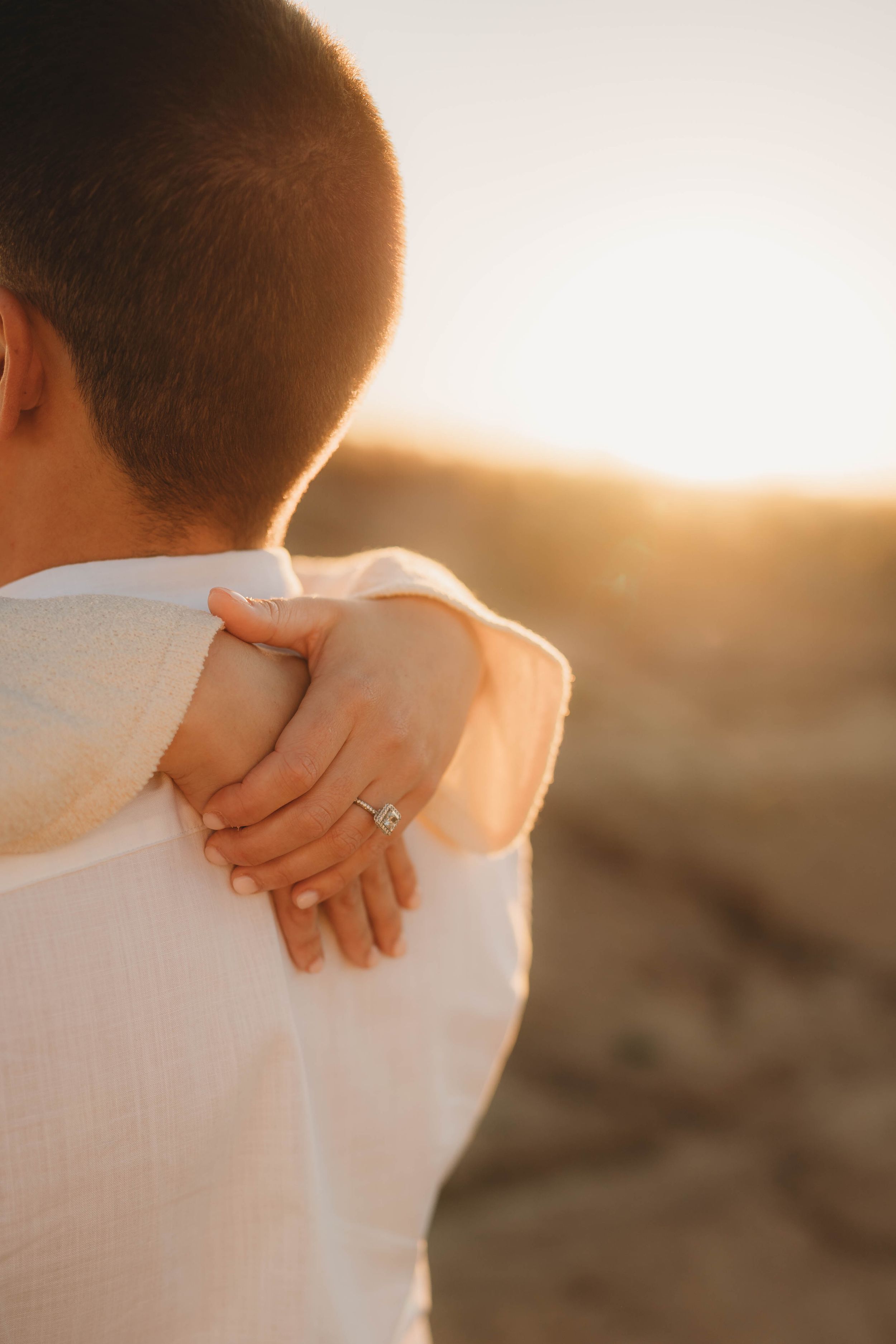 Close-up of womans hand with diamond engagement ring resting on mans shoulder at sunset.