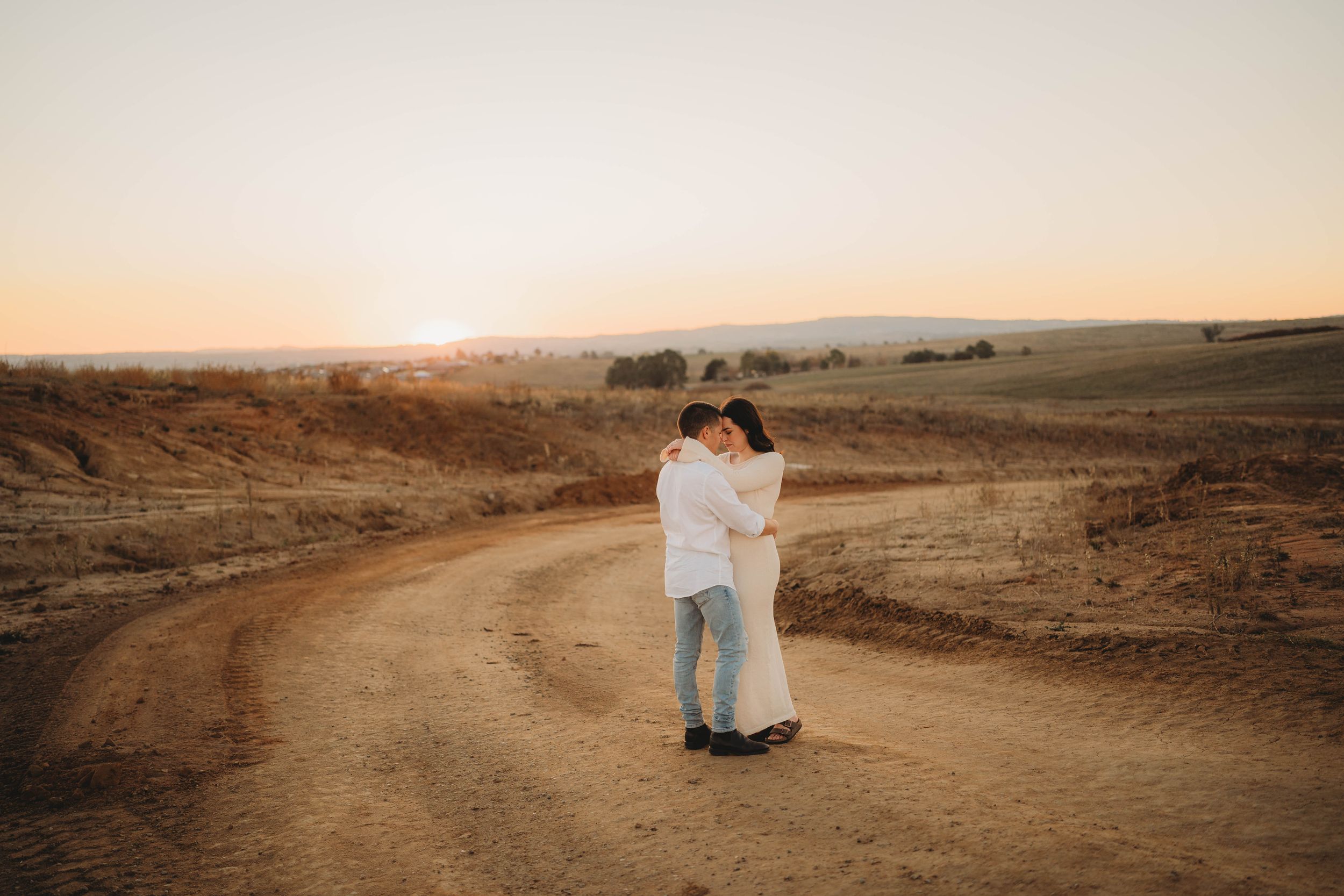 Couple embracing on a winding dirt road surrounded by rolling hills at sunset.