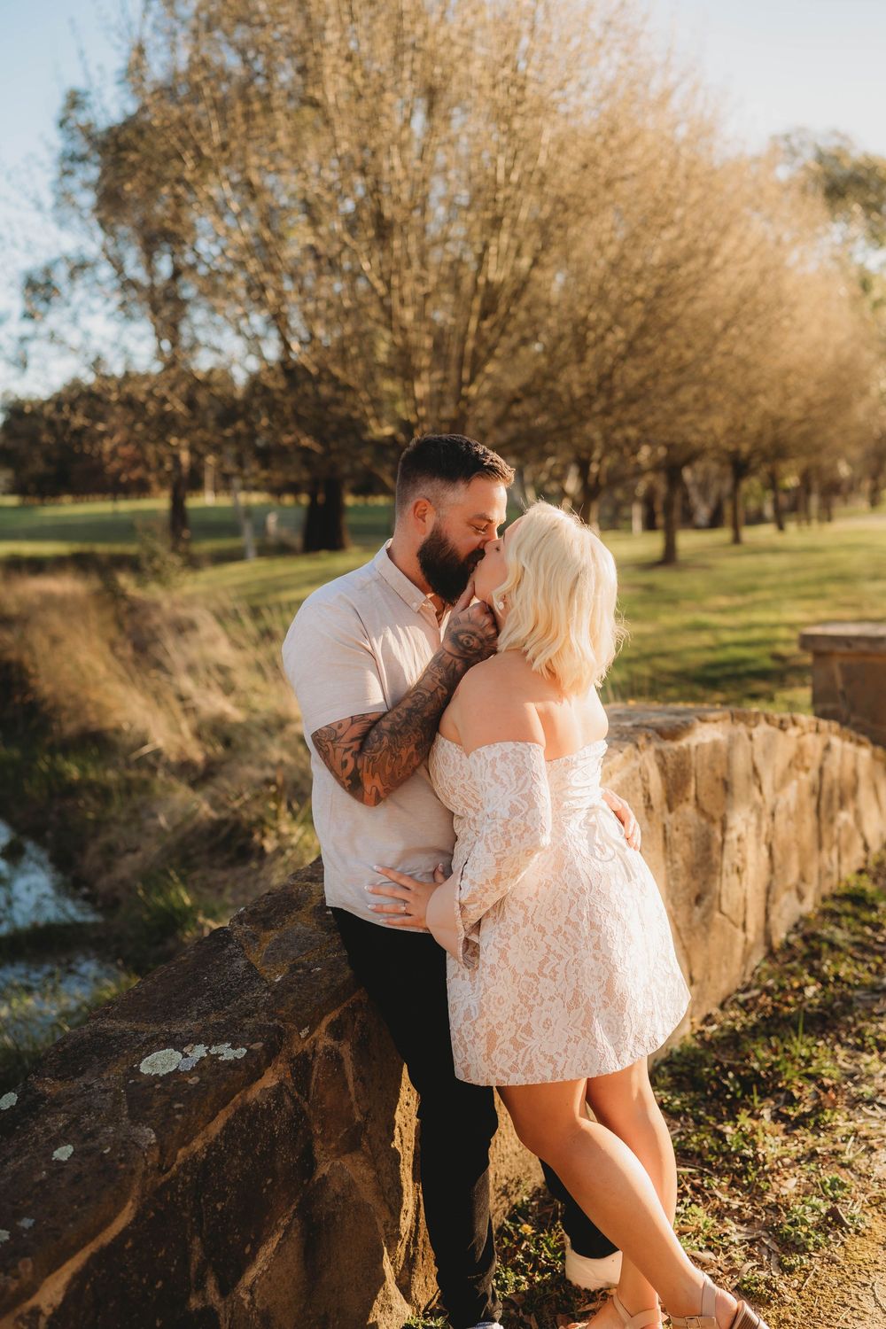 Couple sharing a romantic moment near a stone wall in a sunlit park, man with tattoos embracing blonde woman in lace dress.