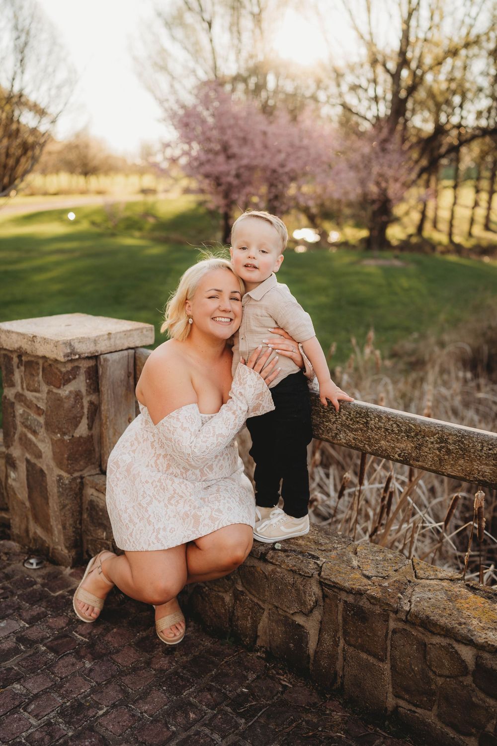 Smiling blonde woman in white lace dress crouching beside toddler standing on stone wall in sunny garden.