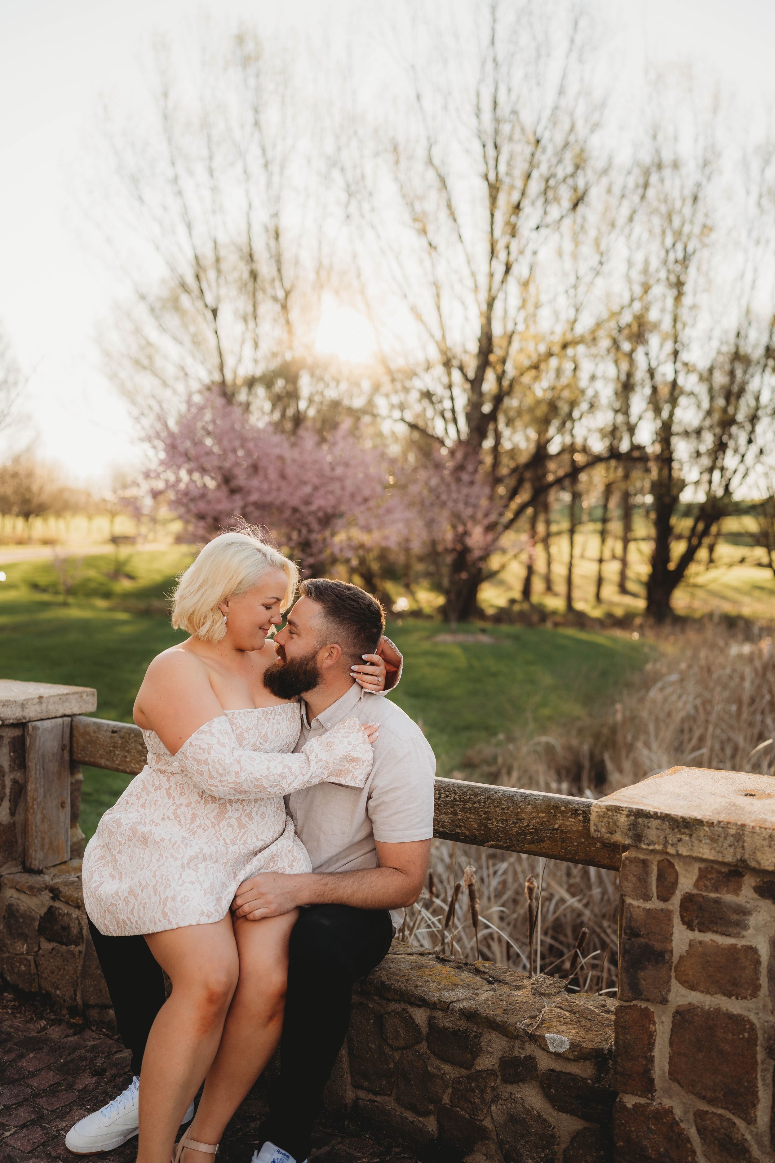 Couple sitting intimately on stone wall at sunset, woman in lace dress embracing bearded man with cherry blossoms behind.