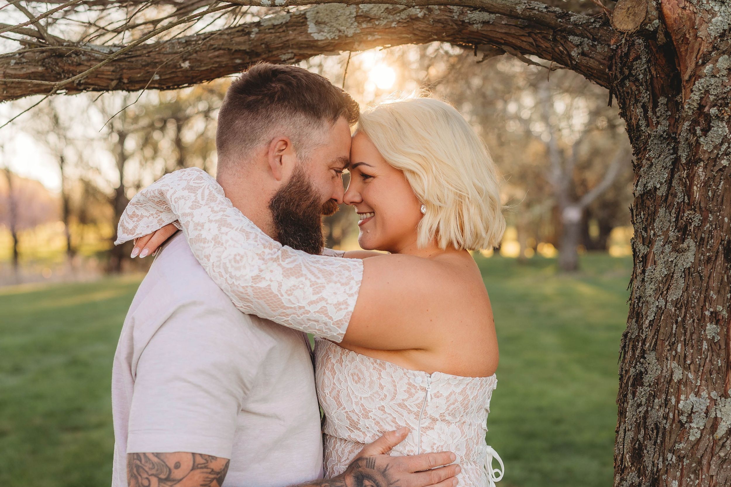 Couple embracing and smiling under a rustic tree branch at golden hour in a lush green park.