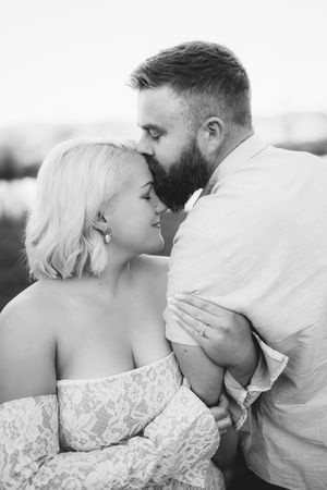 Black and white photo of bearded man kissing forehead of blonde woman in off-shoulder lace dress outdoors.