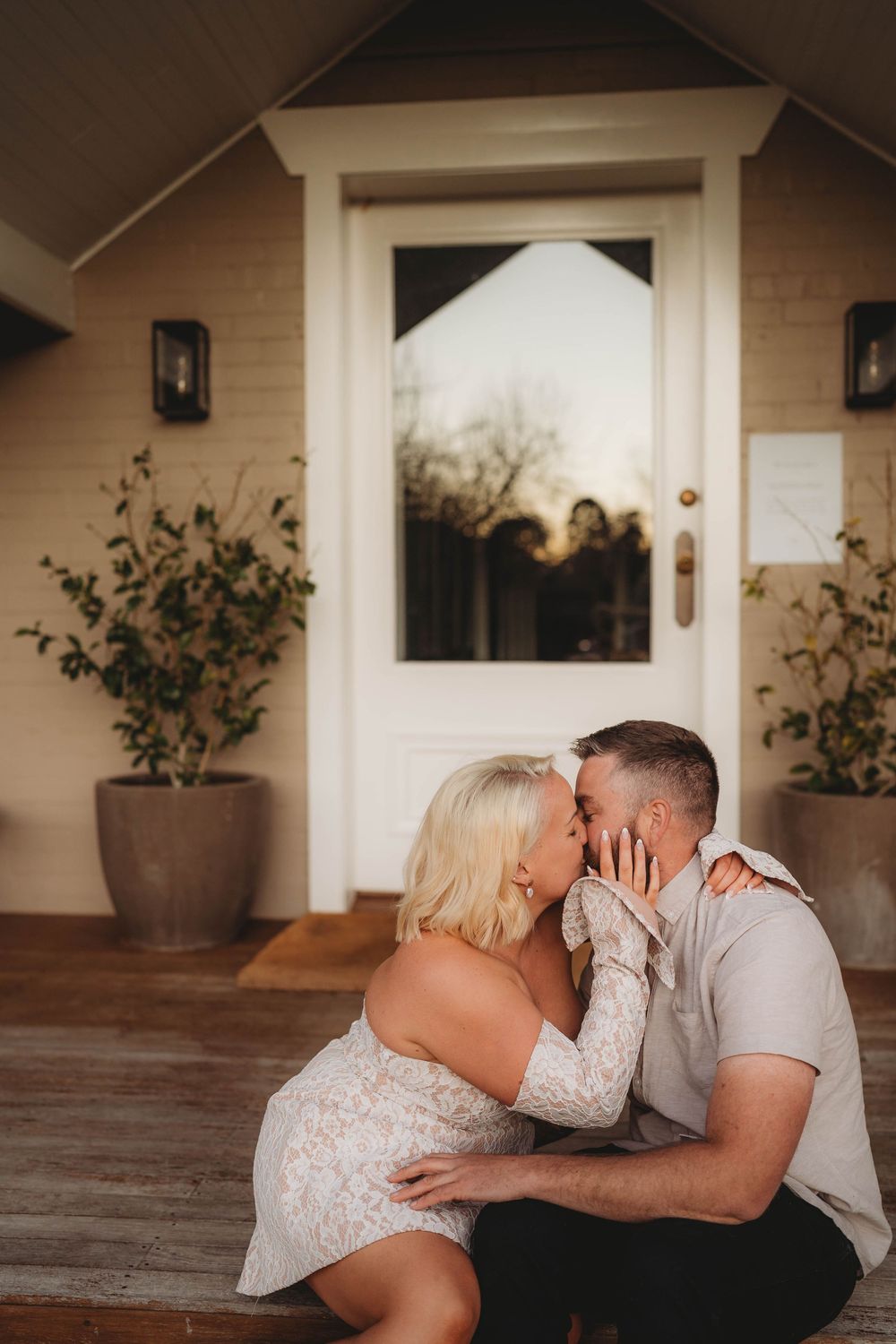 Couple sharing a kiss while seated on wooden porch steps in front of a white door at dusk.