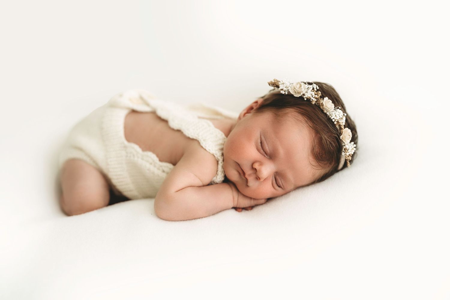 Sleeping newborn baby in cream lace romper and floral headband on white background.