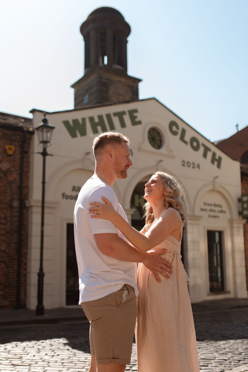 A romantic couple shares a loving moment outside White Cloth building on a sunny day with cobblestone street in frame. Leeds City Centre.