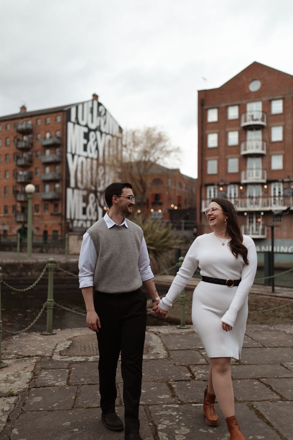 A couple laughs together while walking on a stone path in front of brick apartment buildings on an overcast day.