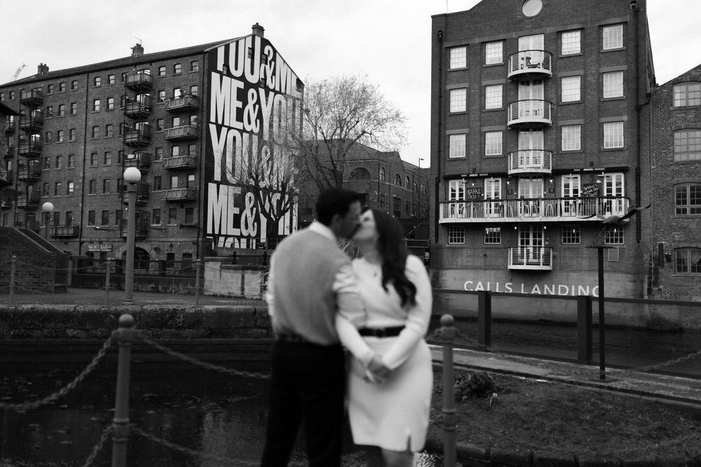 Two figures stand together against an urban backdrop of brick apartment buildings on a river embankment. Leeds City Centre Pre Wedding.