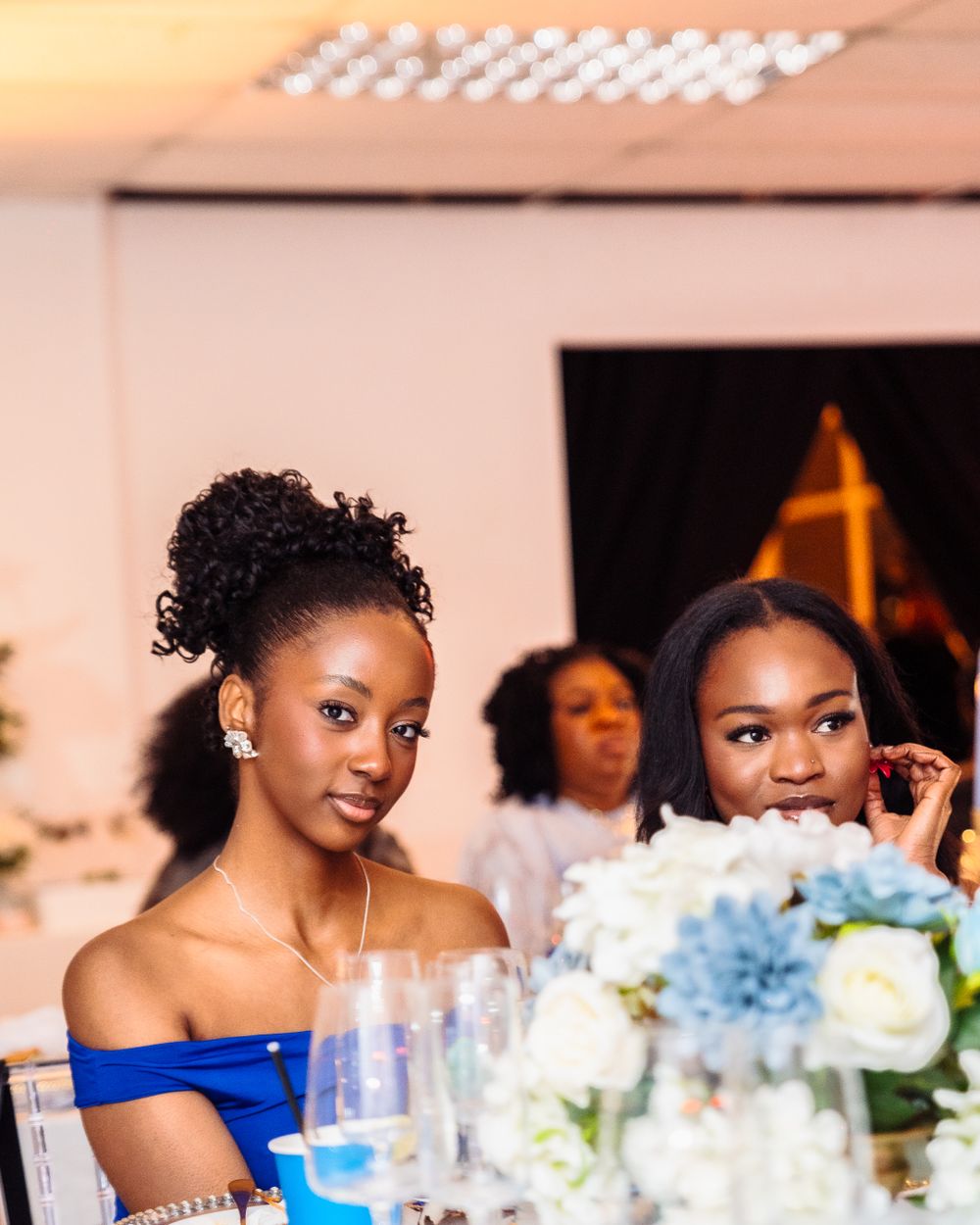 Two elegant women in blue off-shoulder dresses seated at a wedding reception table with floral centerpieces.