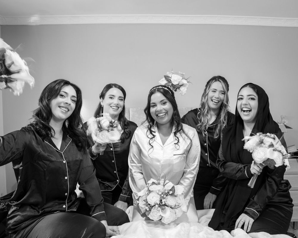 Black and white photo of bride in white robe laughing with four bridesmaids in black pajamas on a bed.