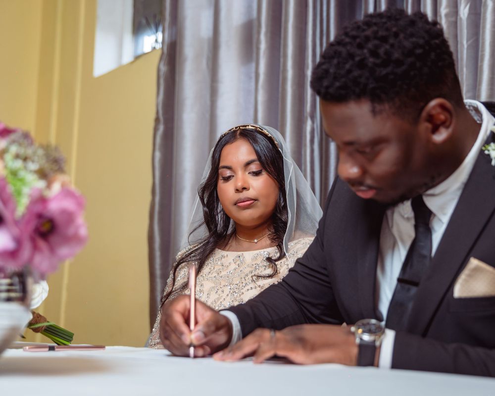 Bride in lace veil watches as groom signs wedding documents at a ceremony table with pink flowers.
