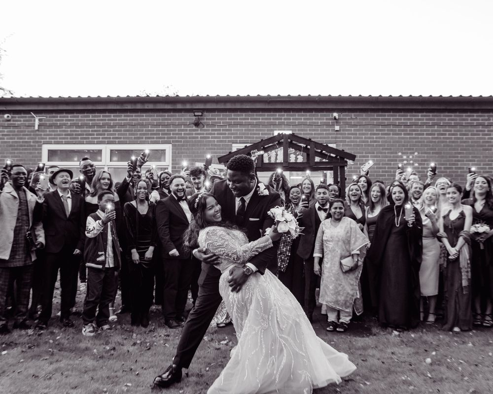 Black and white photo of newlyweds dancing as guests celebrate around them outside a brick building.