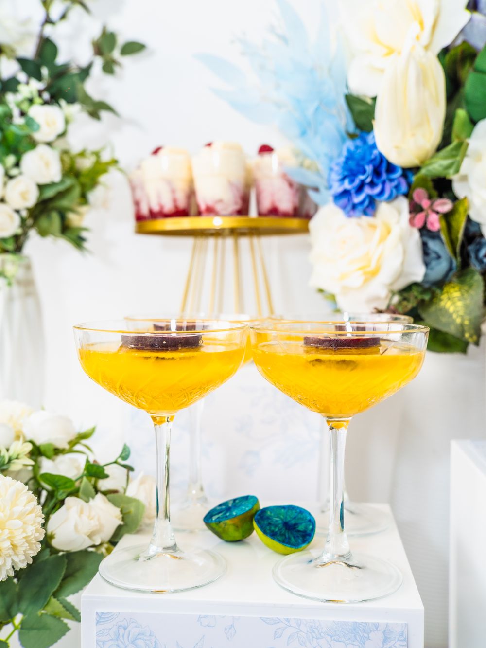 Two golden cocktails in coupe glasses with blue floral arrangements and desserts at a wedding reception.
