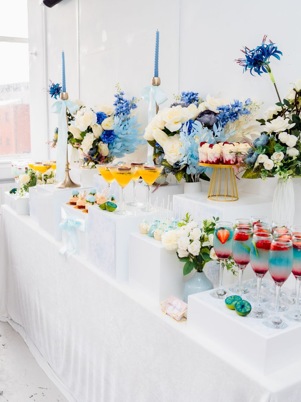 Elegant wedding reception dessert table with colorful cocktails, blue candles, and white and blue floral arrangements.