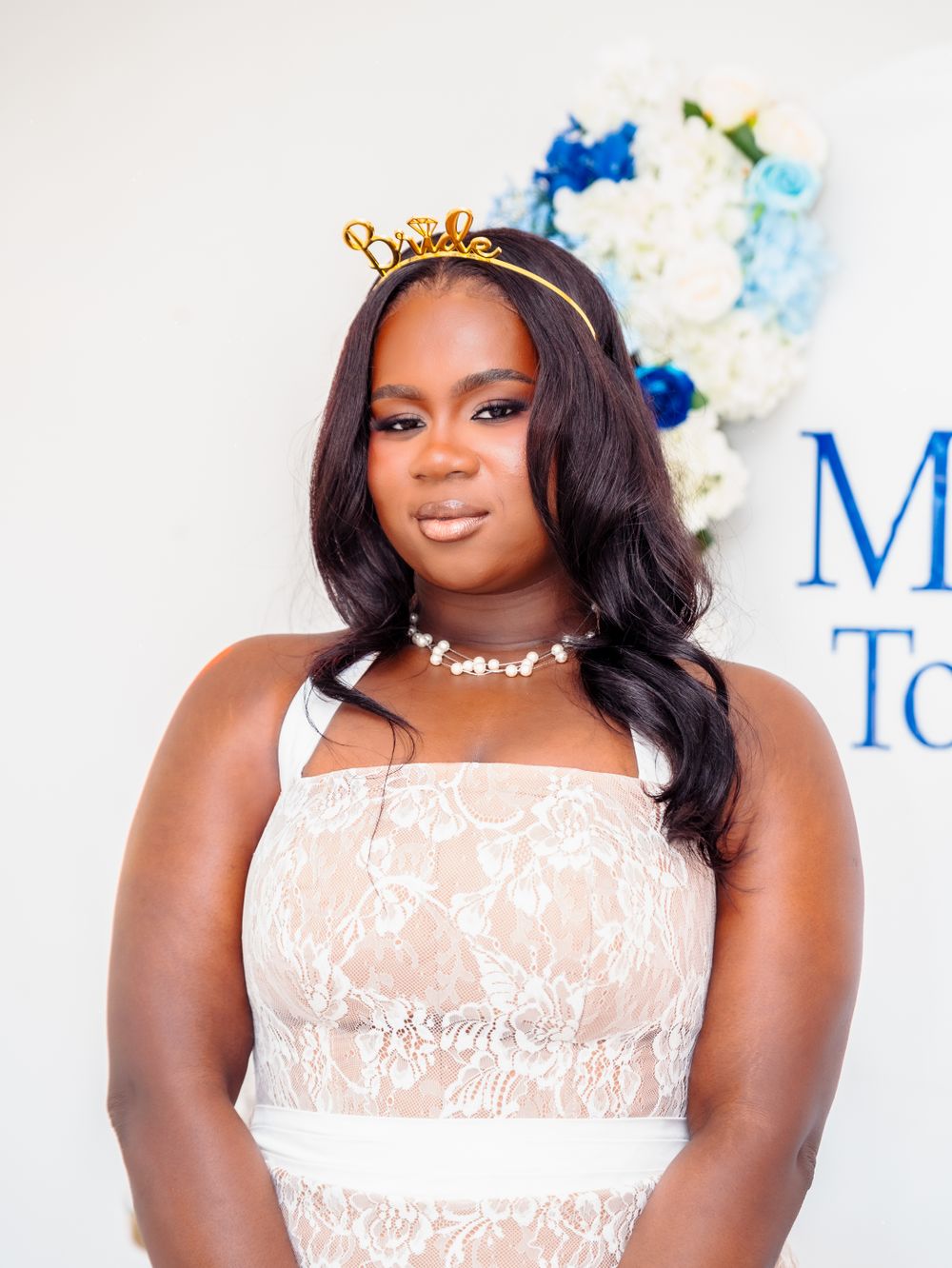Bride wearing a gold crown headband and white lace dress posing in front of a floral backdrop.