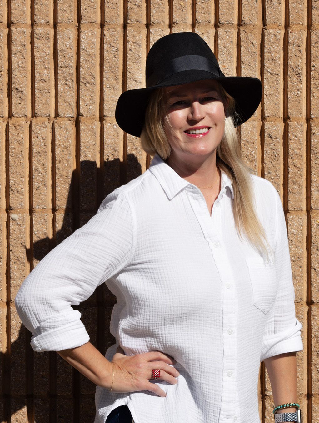 Confident female luxury photographer in white shirt and black hat, posing in sunlight against a modern tan brick wall.