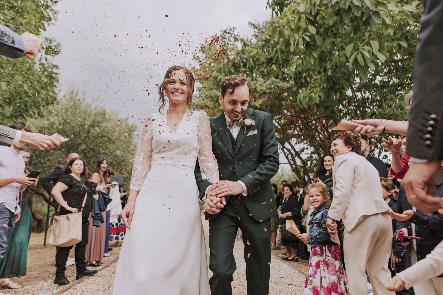 Newlyweds walk down a path surrounded by guests throwing confetti during an outdoor wedding celebration.