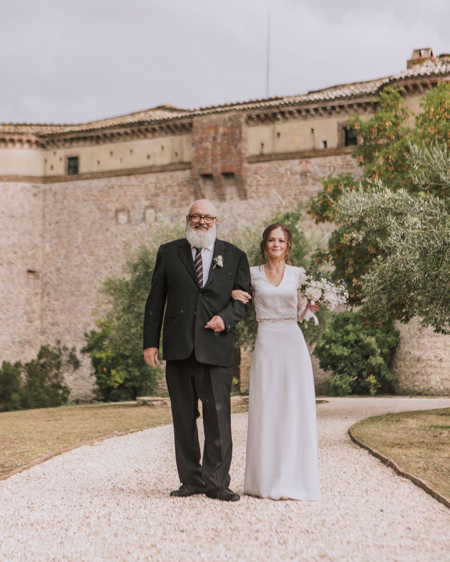 Wedding couple poses in front of historic stone fortress building on gravel path.