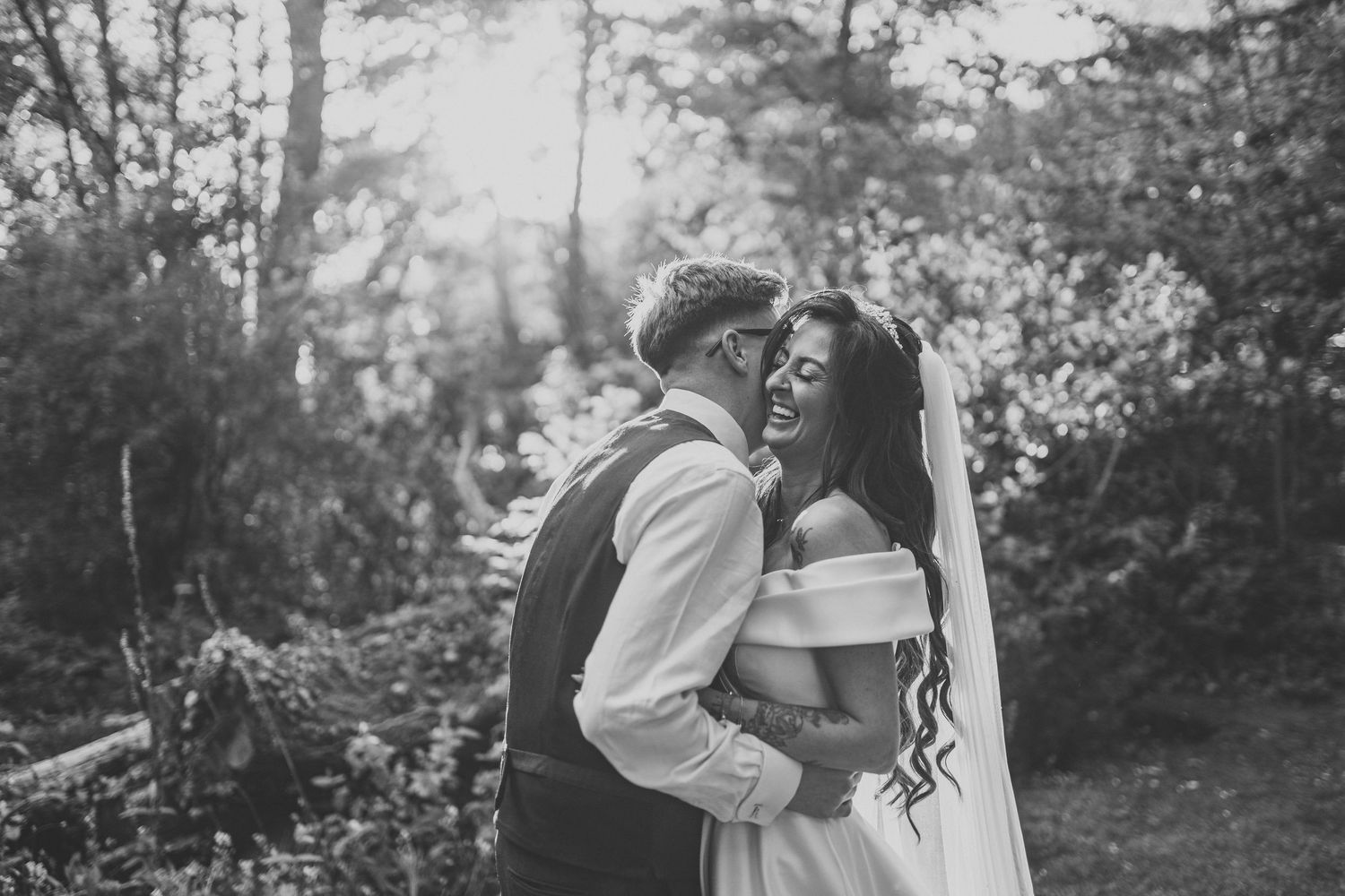 Black and white photo of a bride and groom embracing and laughing together outdoors among lush trees at Brook Hall Hotel, Ellesmere Port
