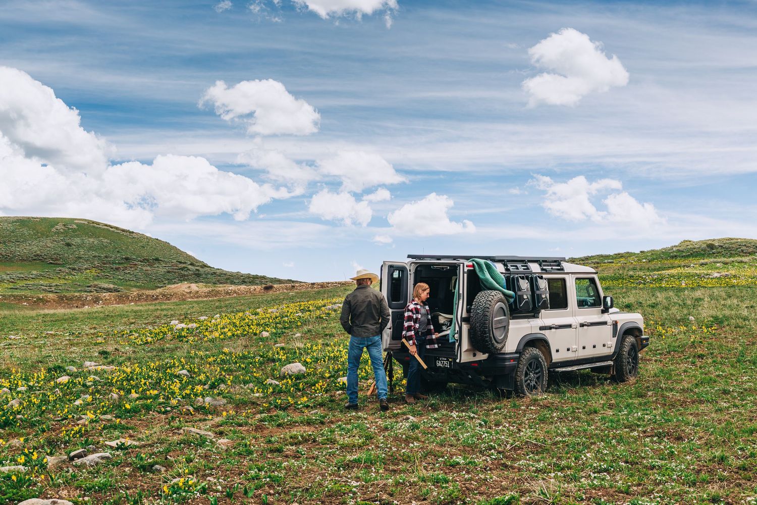 Two people standing beside a white Land Rover Defender in a scenic green meadow under a cloudy sky.