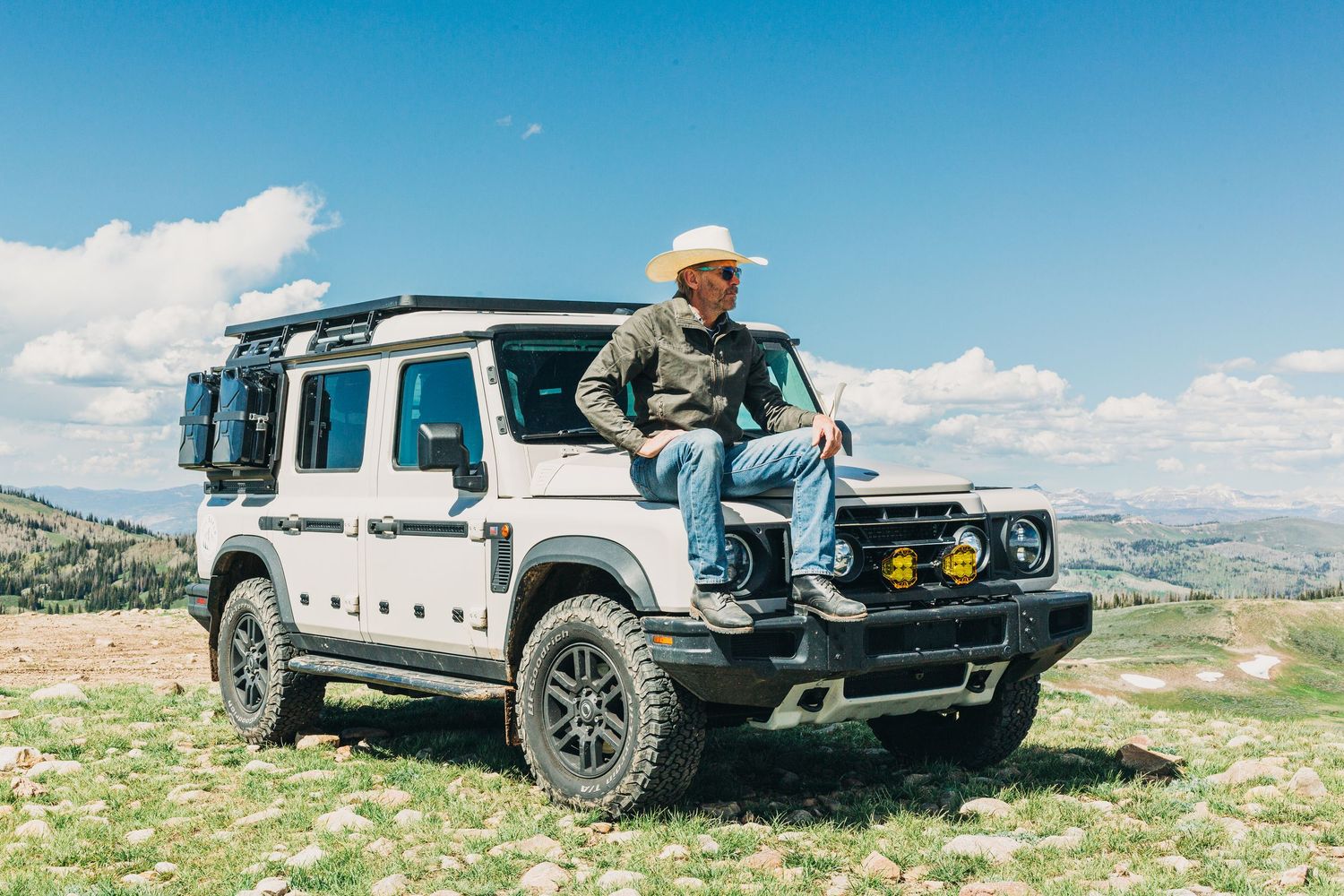 Man in white cowboy hat sitting on hood of white Land Rover Defender on a hilltop with panoramic views.