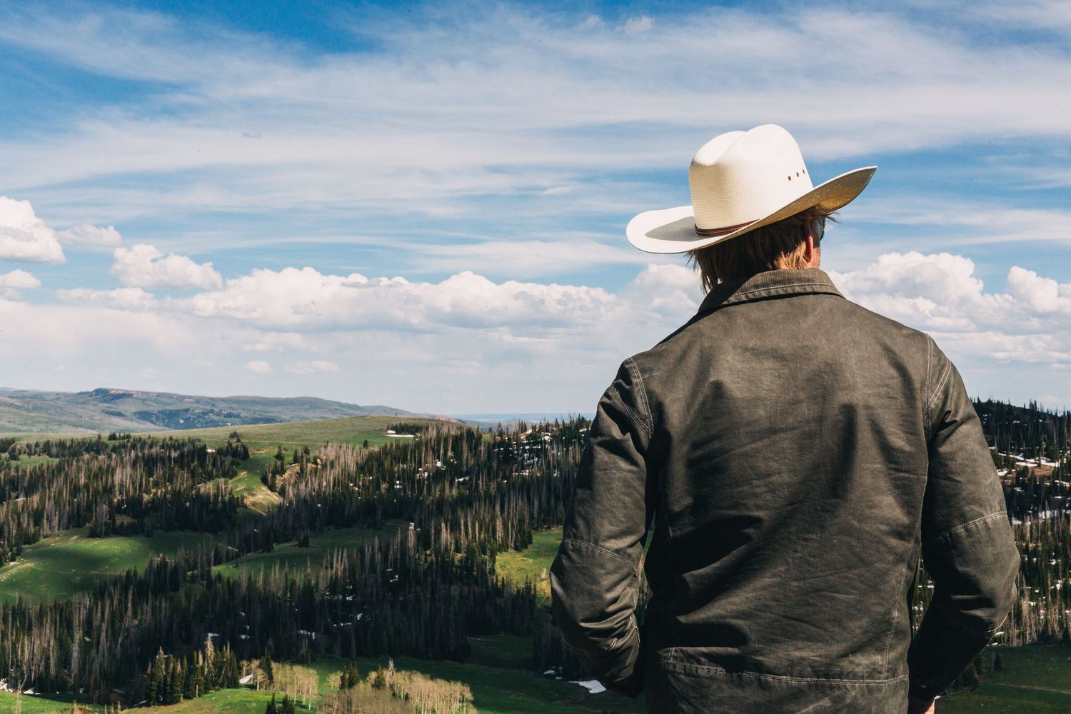 Man in white cowboy hat and jacket overlooking a lush green valley with trees under a blue sky.