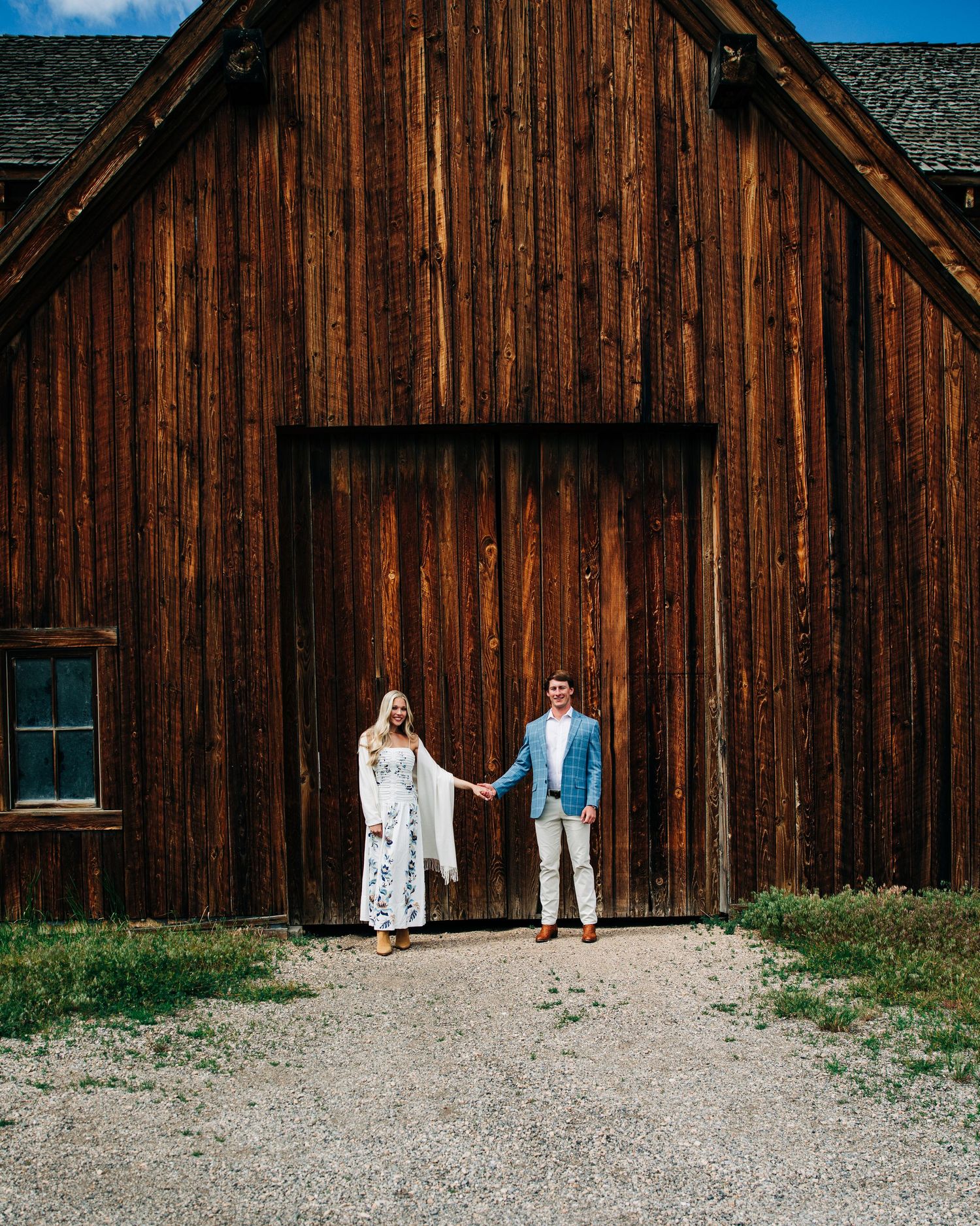 Couple holding hands in front of rustic wooden barn, woman in white lace dress, man in light blue suit, outdoor setting.