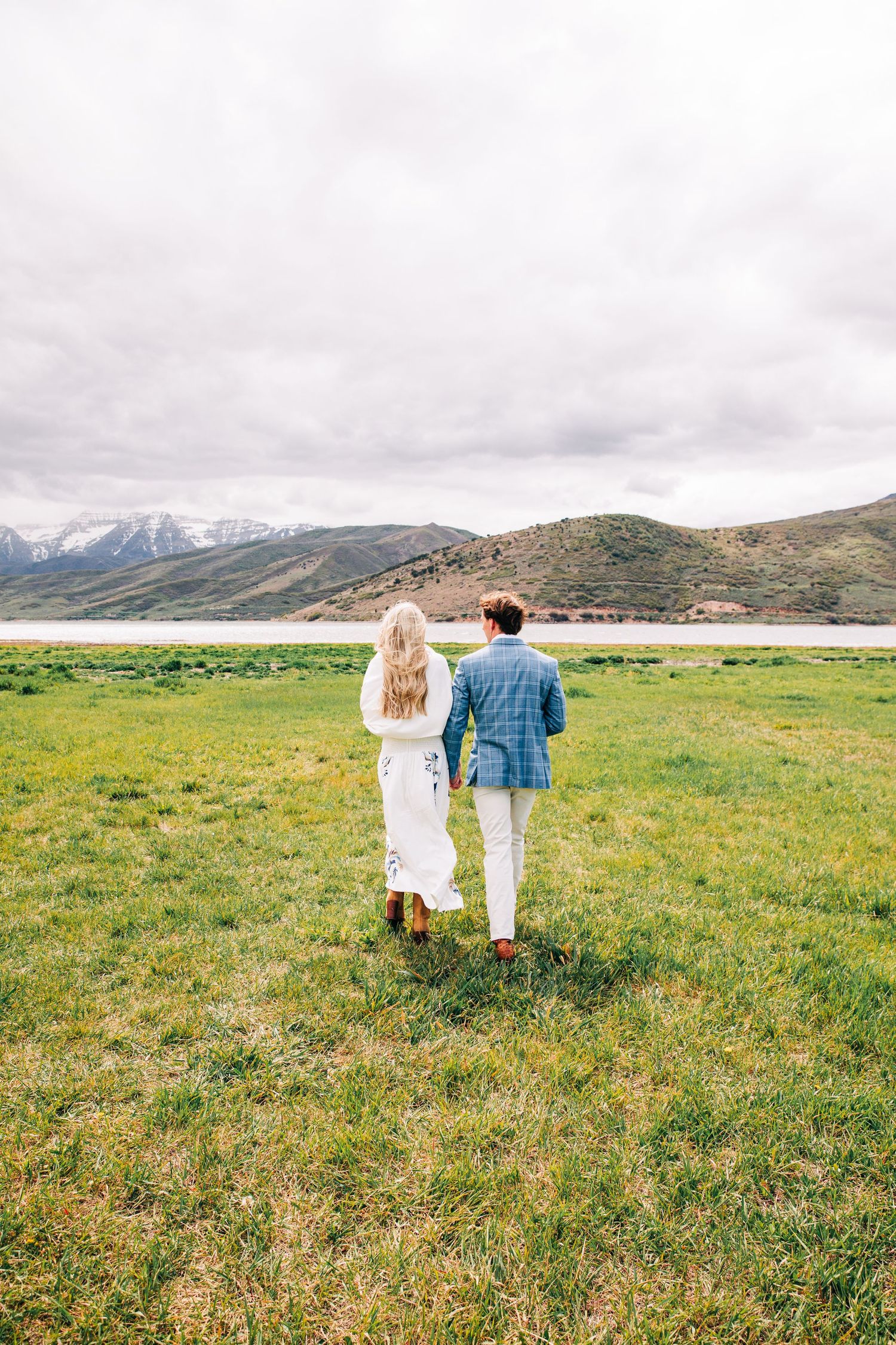 Couple walking hand in hand through green meadow with mountain lake backdrop on cloudy day.