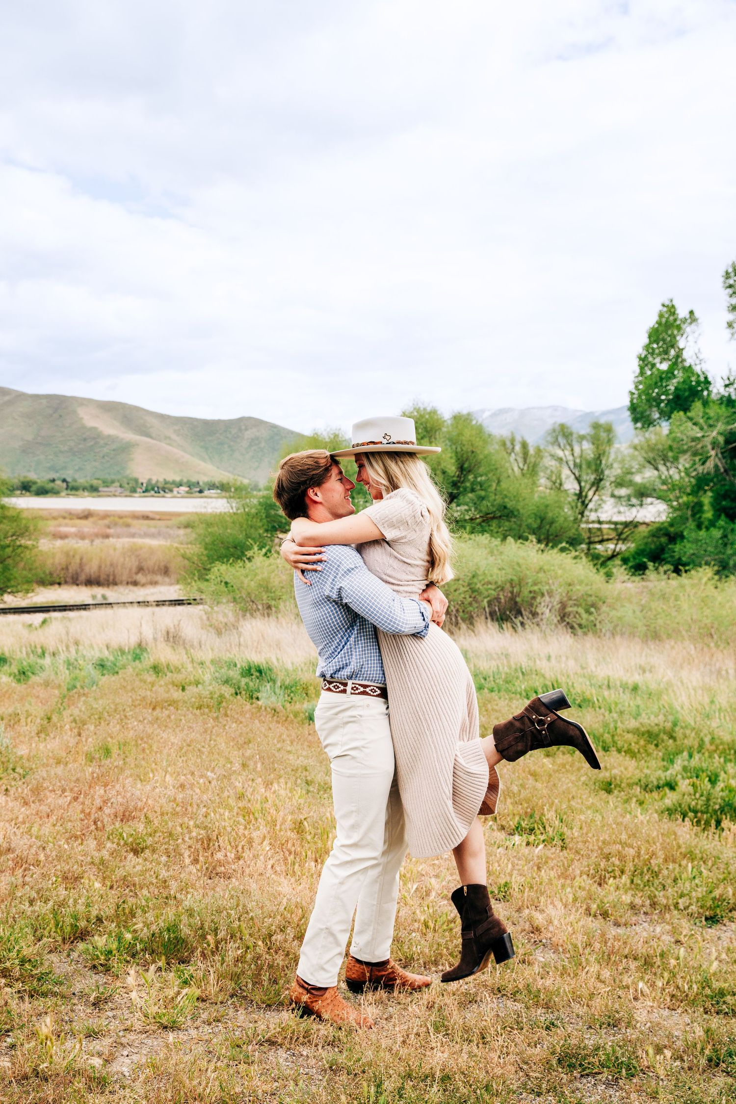 Couple embracing in a field with mountain backdrop, woman in hat and dress lifted by man in white pants.