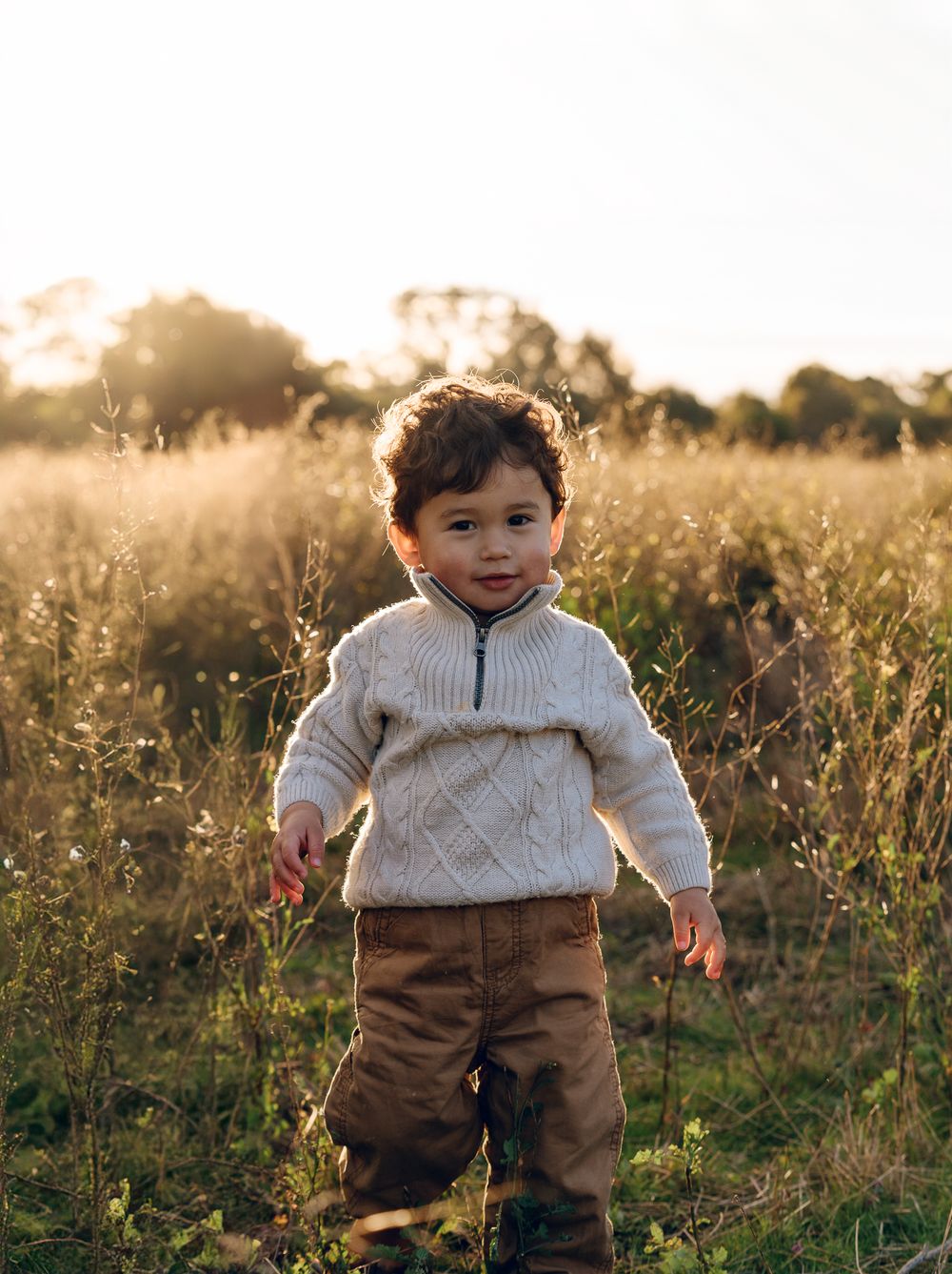 A small child wearing a cream sweater and brown pants plays in a sunlit field at golden hour.