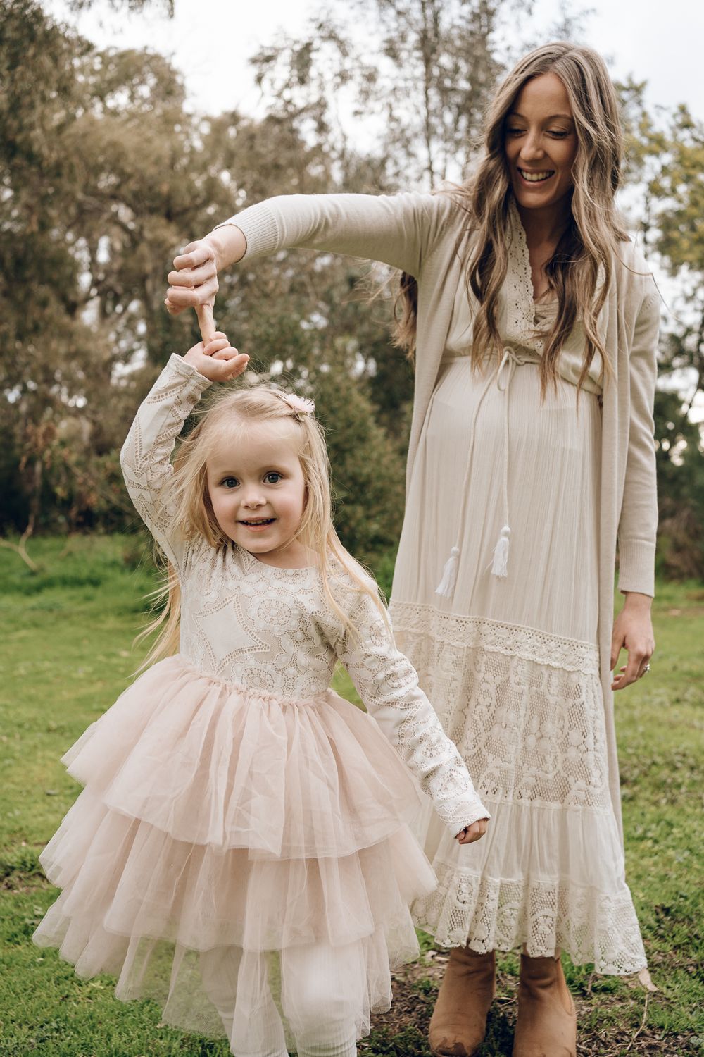 Mother and daughter in matching light colored dresses dance together on green grass.