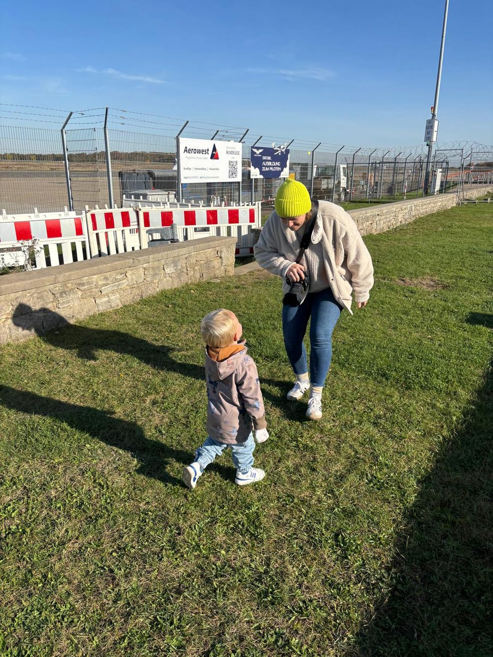 Fotografin spielt mit einem Kind am Braunschweiger Flughafen und macht dabei Familienfotos, die Sonne scheint, sie trägt eine gelbe Mütze.