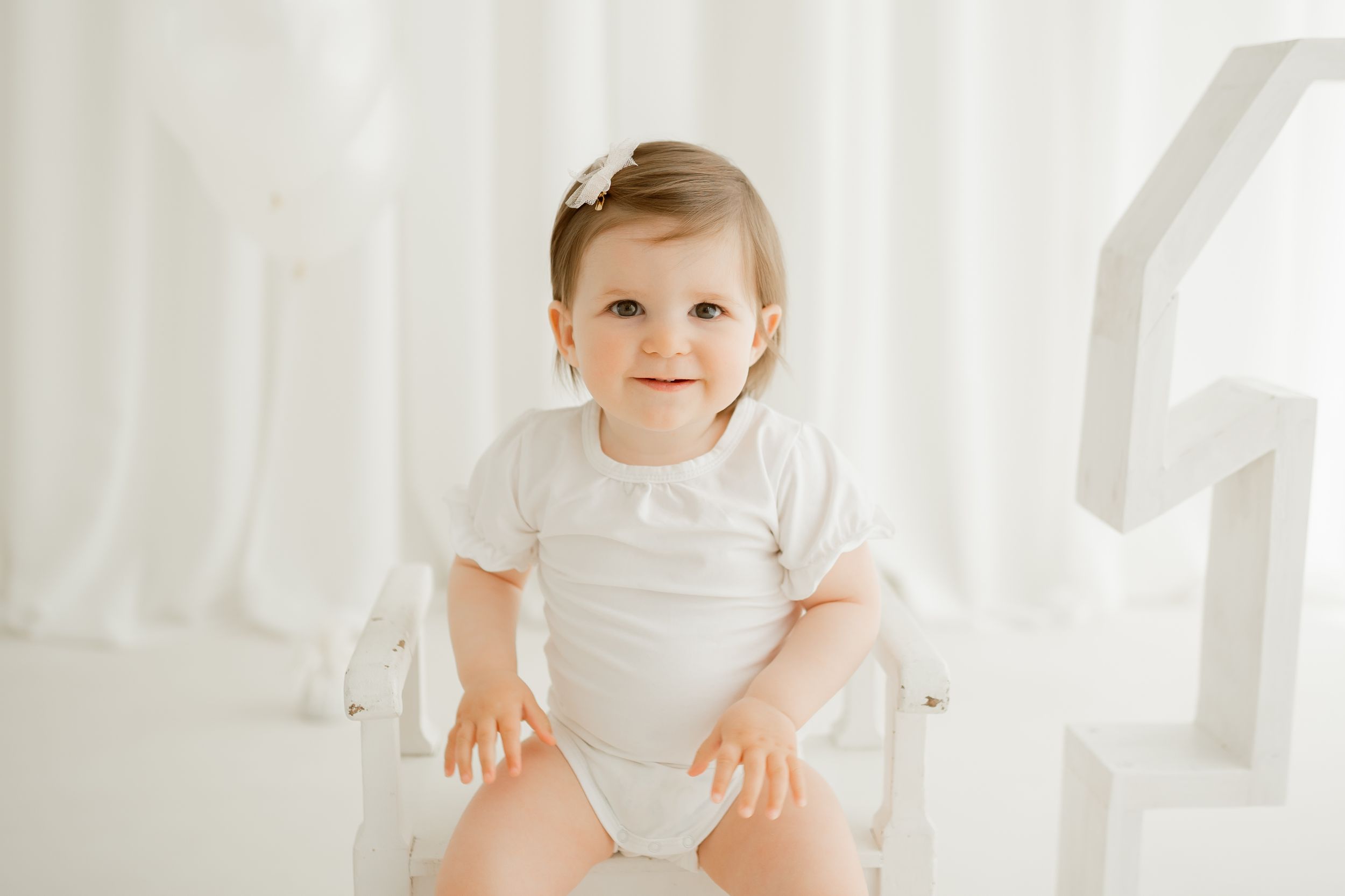 Baby in white outfit sitting in bright white chair against white curtains.