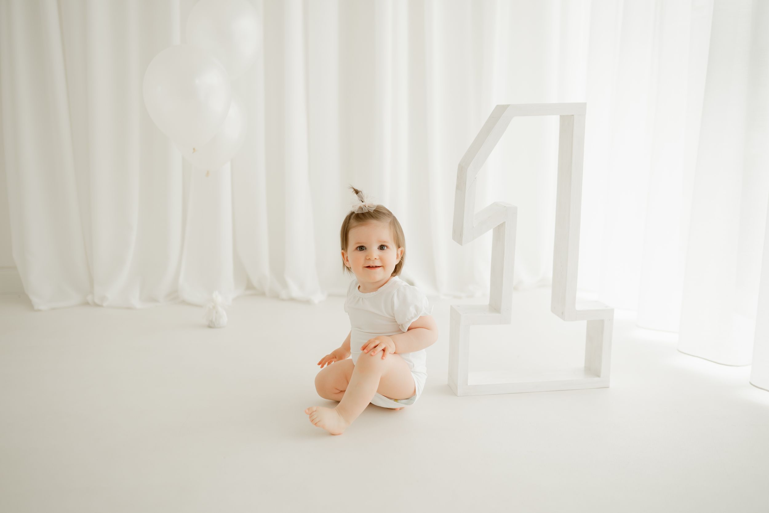 Baby sitting next to large number one decoration in all-white studio setting.