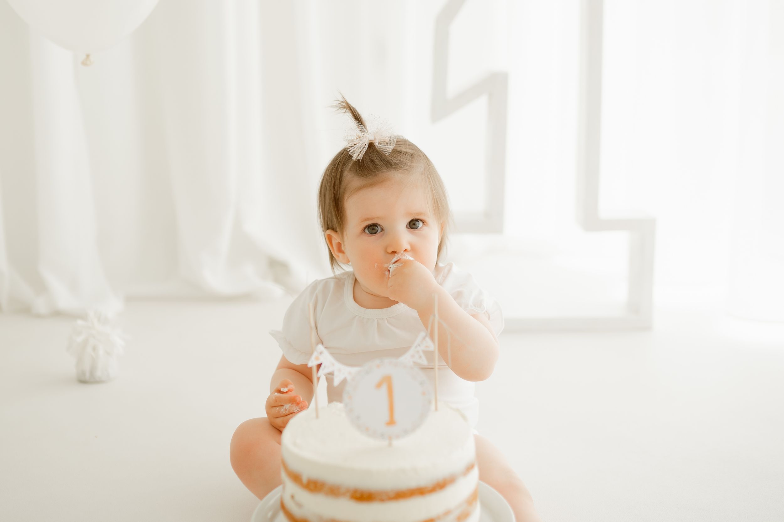 Baby in white dress tasting their first birthday cake with number one candle on top.