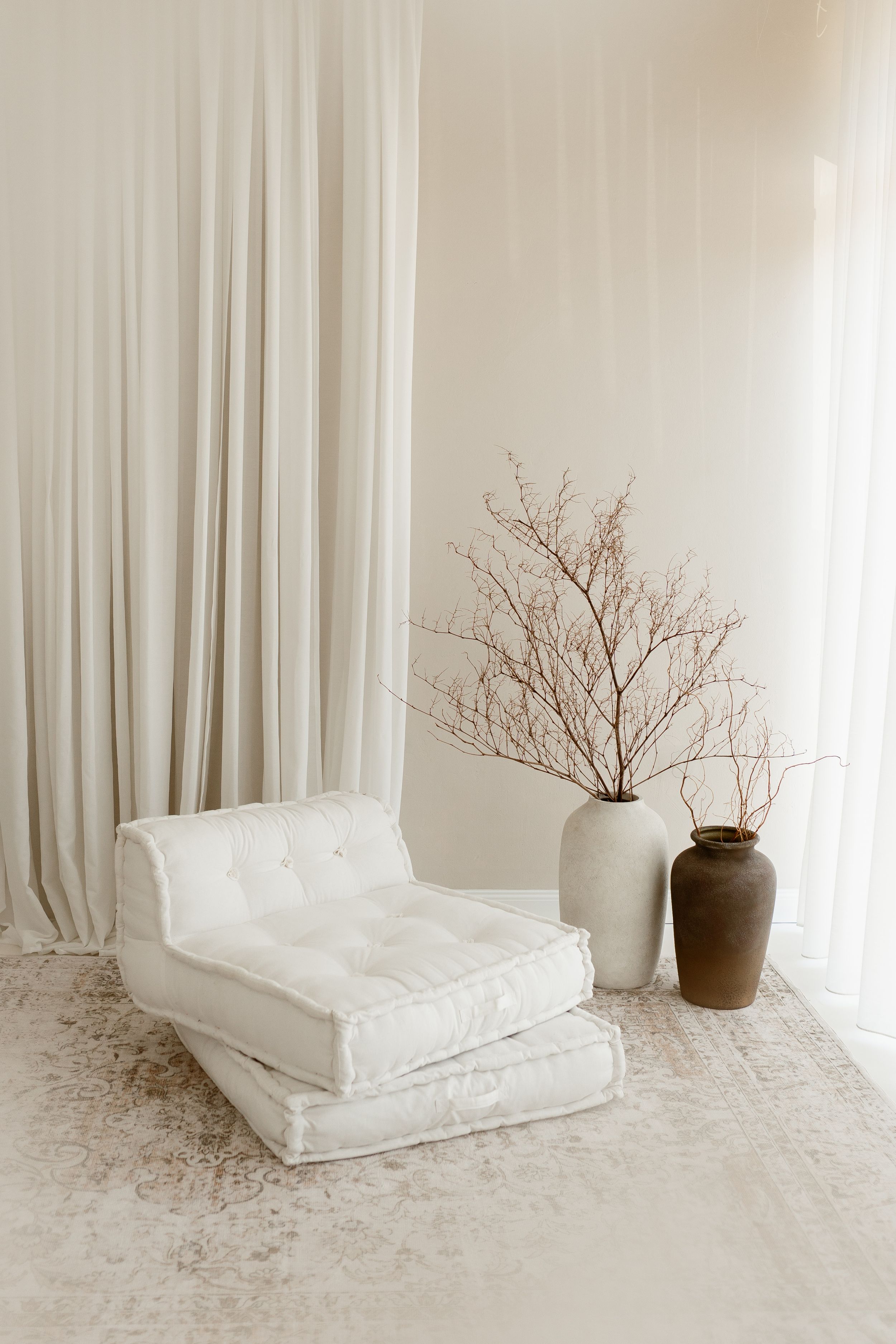 Minimalist white room with tufted floor cushion, sheer curtains, and two vases with dried branches.