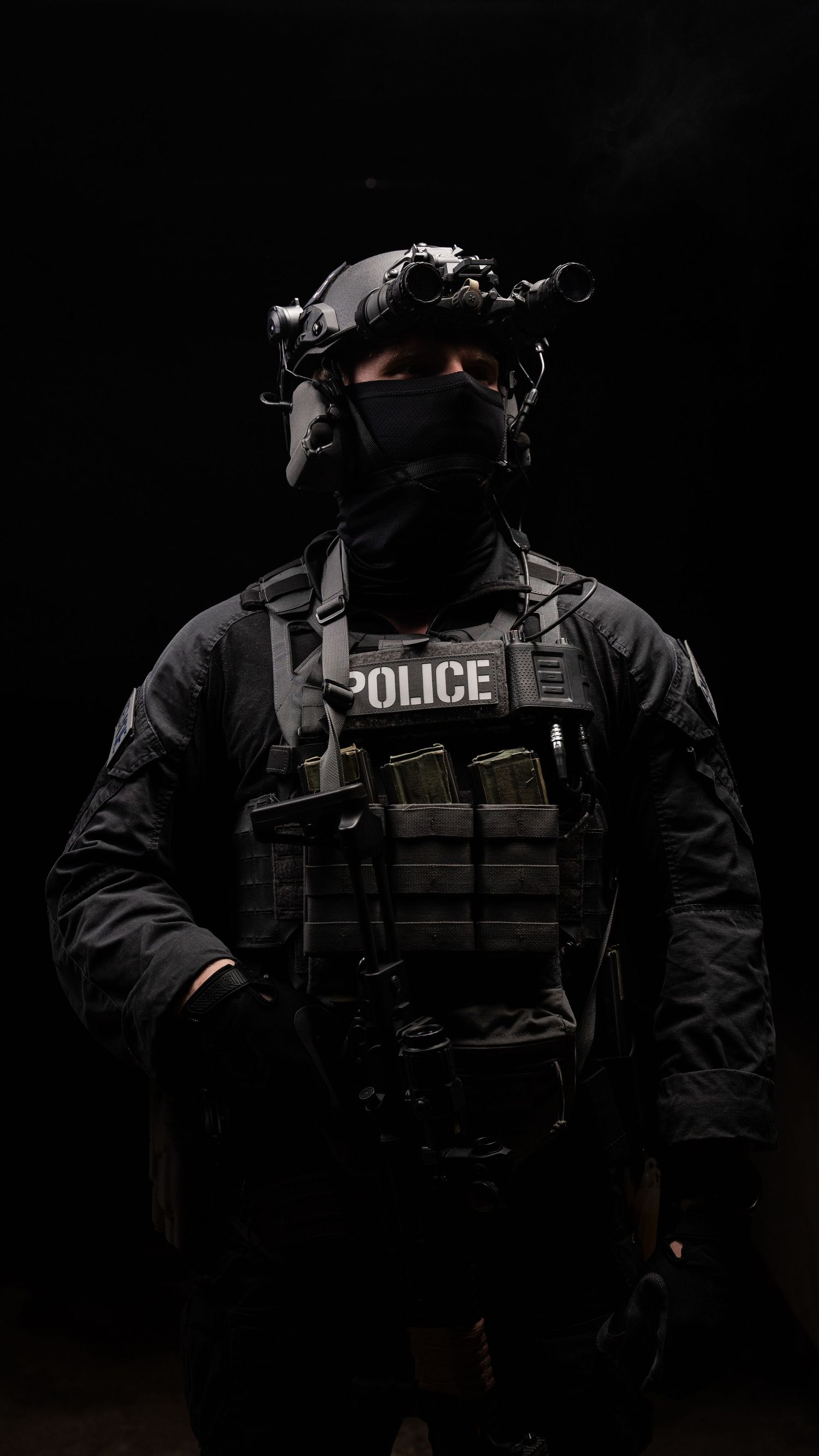 A police officer in tactical gear stands in darkness with night vision equipment against a black background.