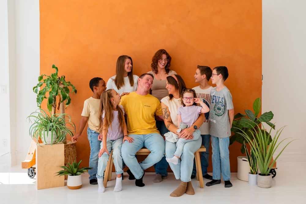 Group photo session with people sitting casually against orange wall with greenery.
