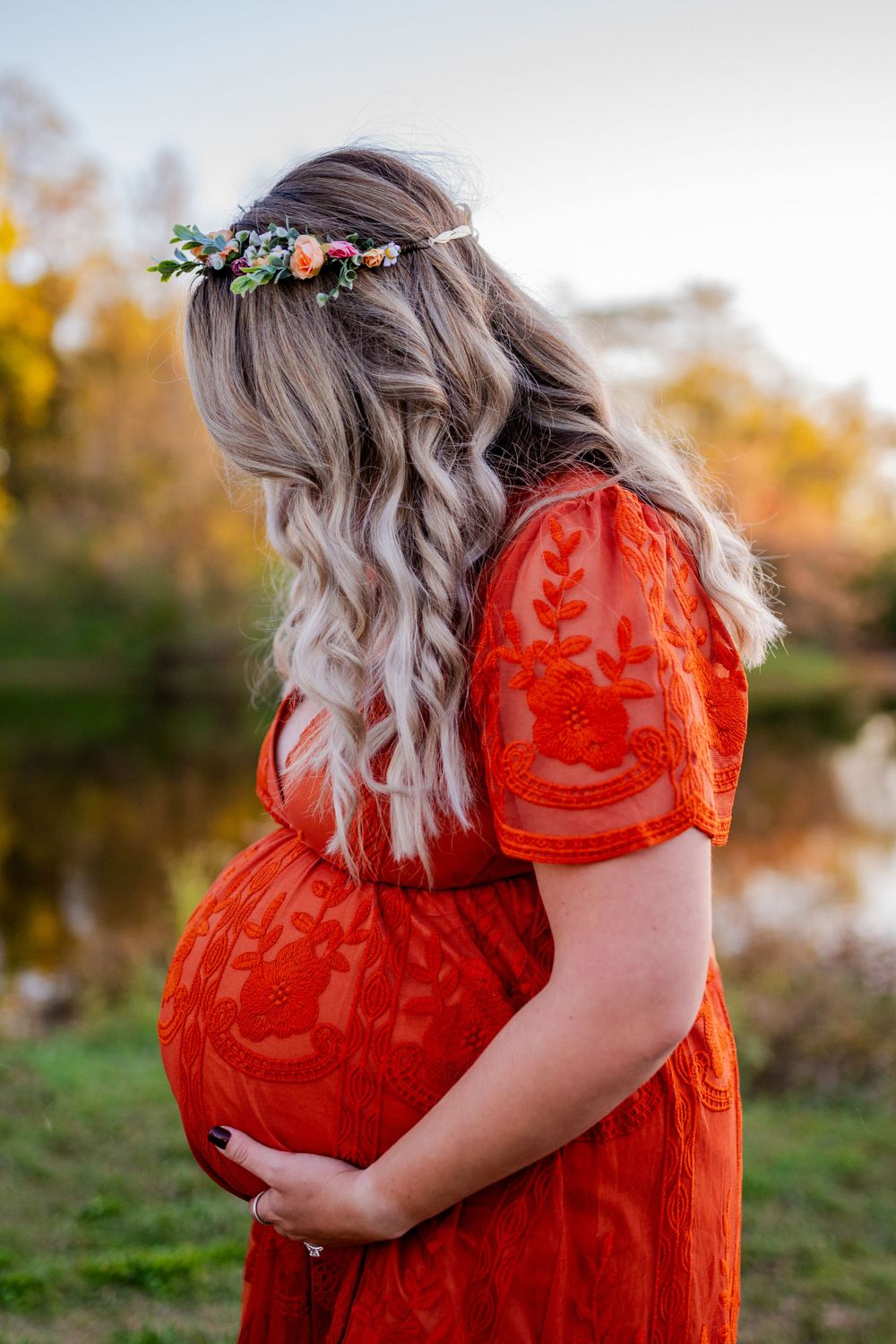 Maternity photos showing a person in a red lace dress by a pond during golden hour.
