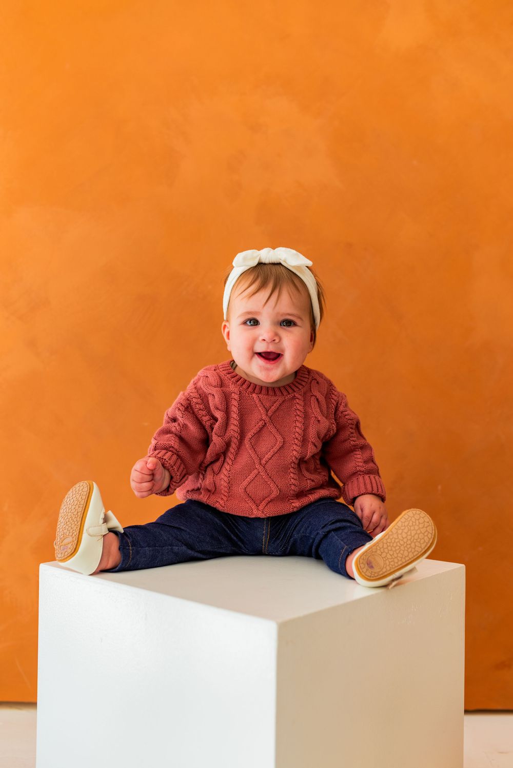 Baby in red sweater sits happily on white platform against orange background.