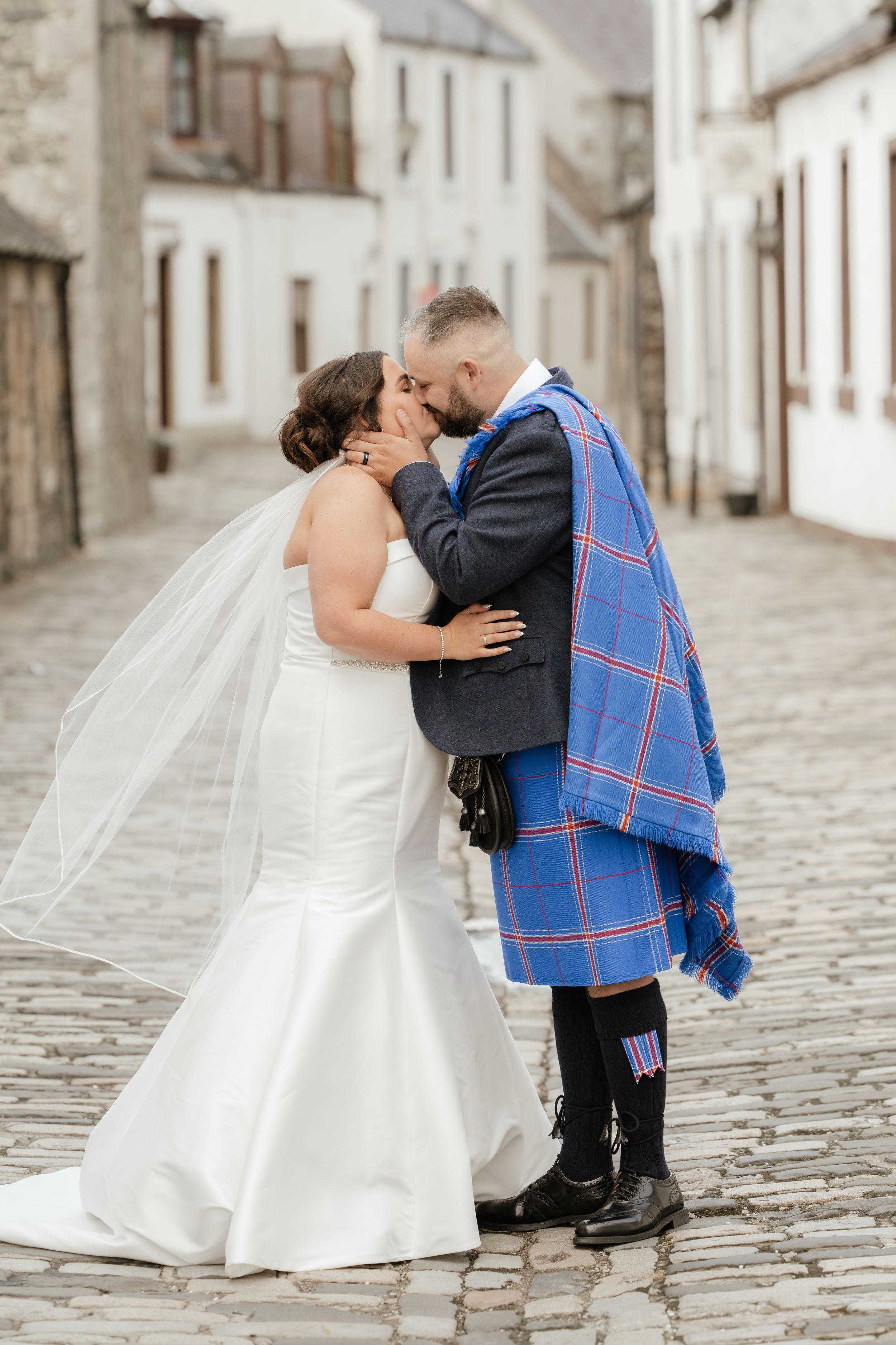 A romantic wedding kiss captured on a cobblestone street in a charming European town with white stucco buildings.