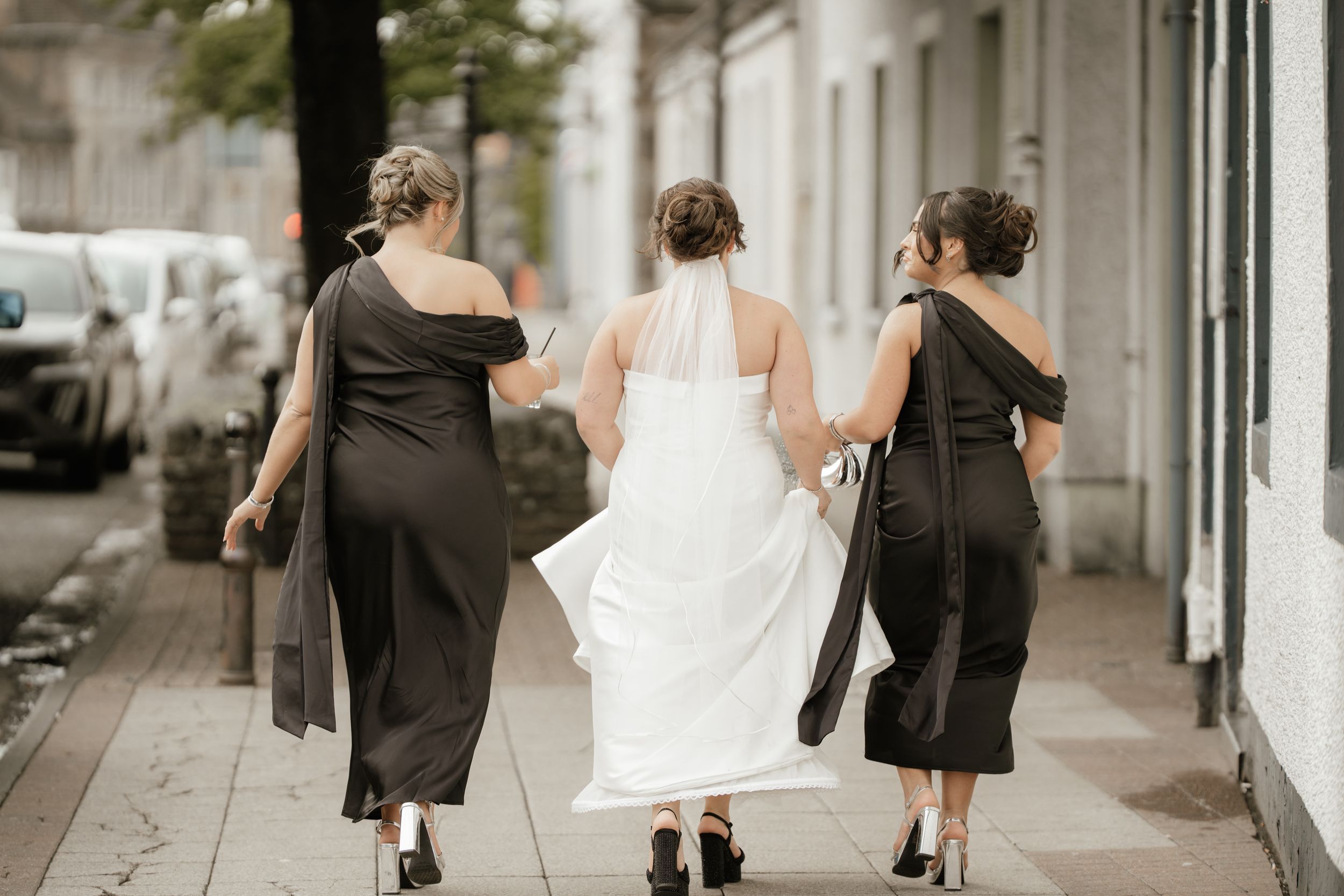 Three people in formal attire walk down a sidewalk, with the center person wearing a flowing white dress.
