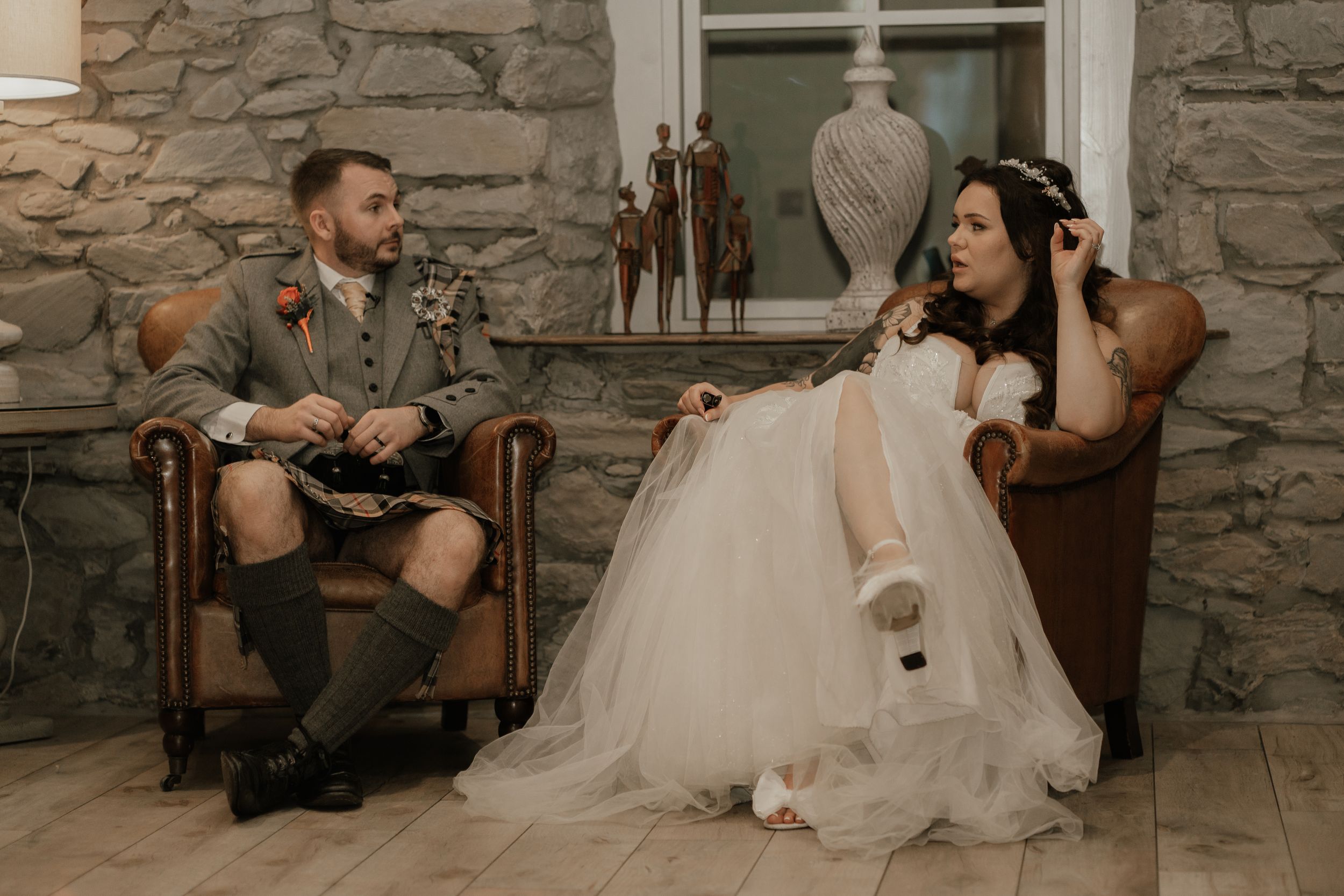 A couple in wedding attire relaxes in leather chairs against a stone wall, with a rustic elegant atmosphere.