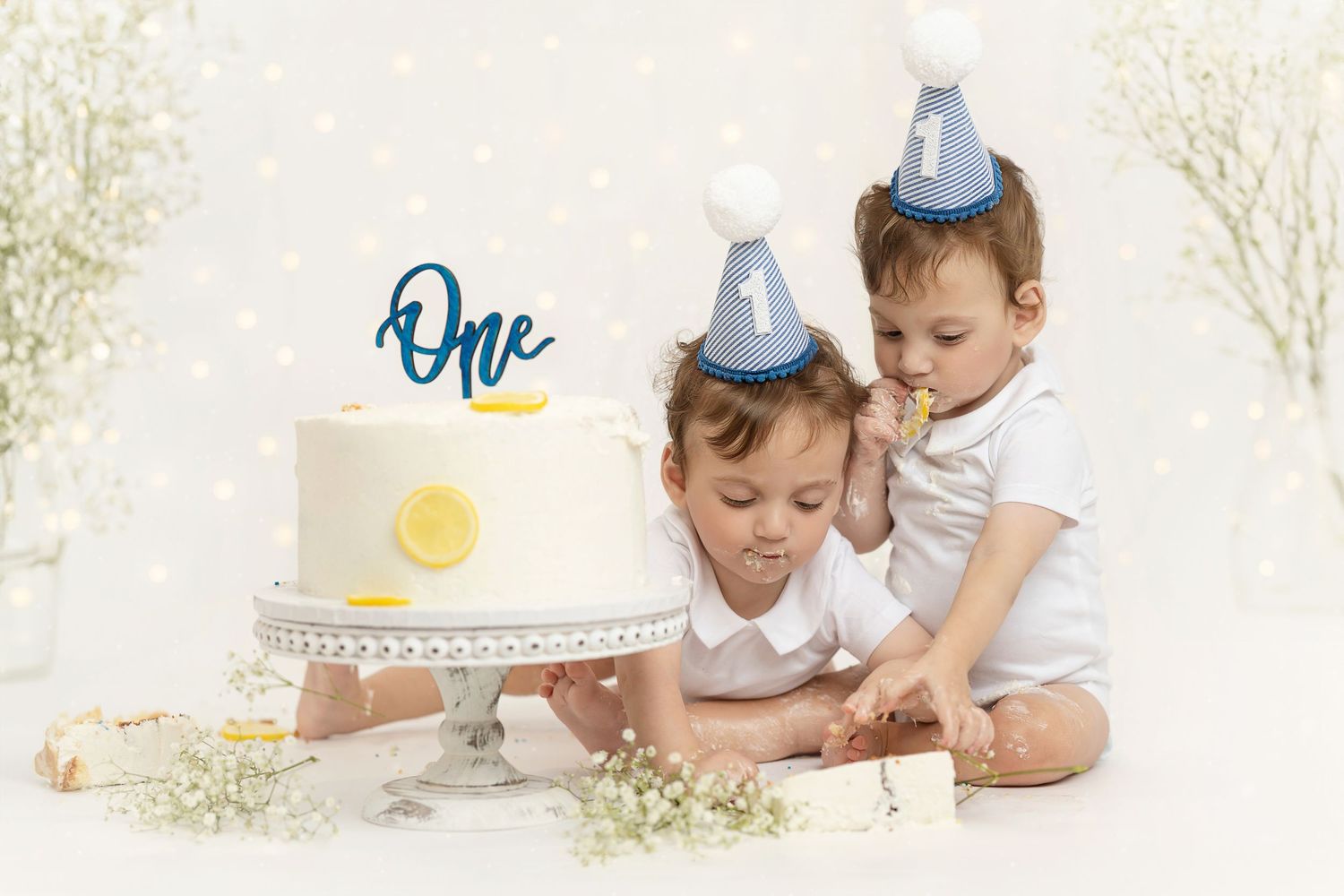 Two children sitting together with teddy bear under decorative hoop on cream backdrop.