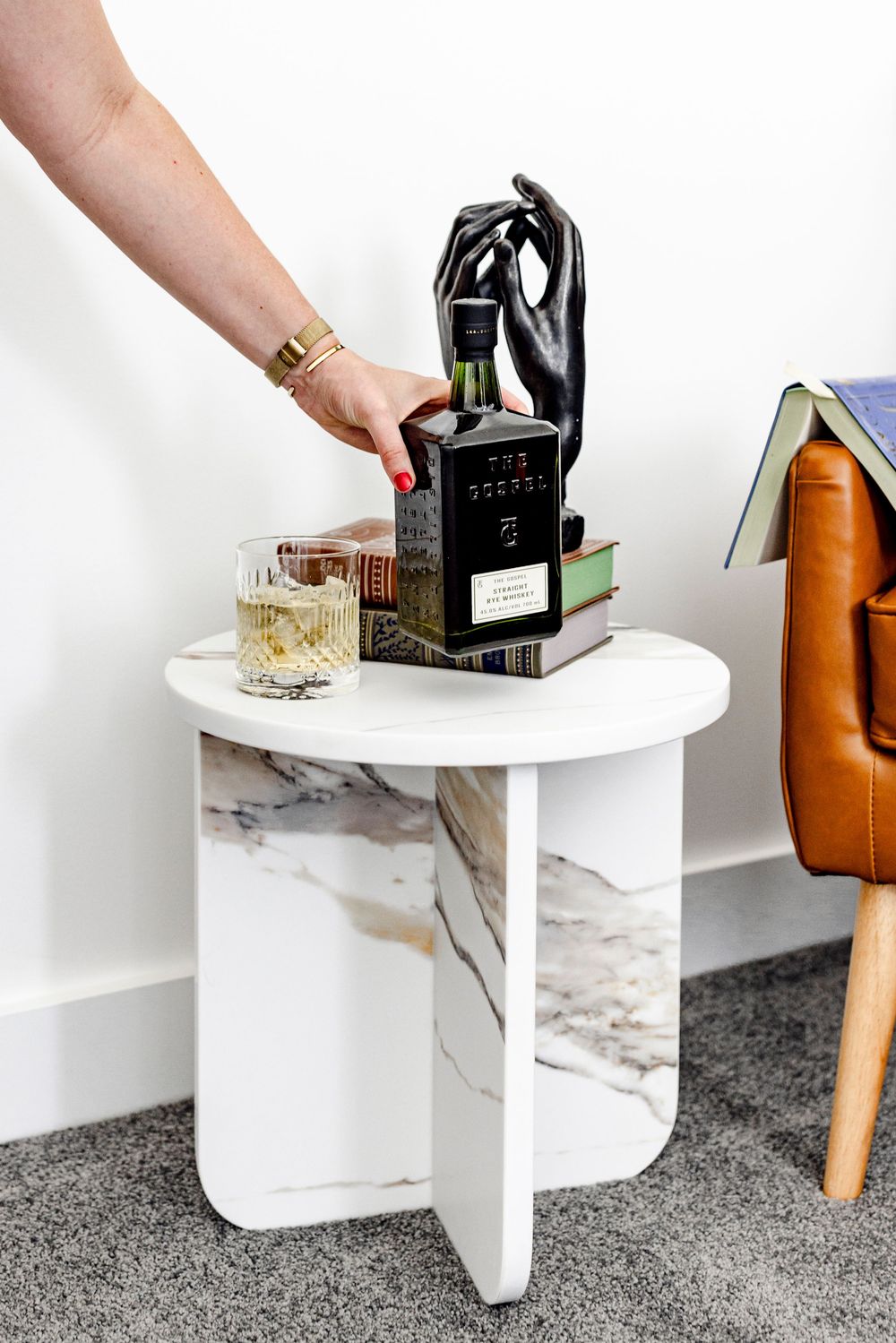 White marble side table styled with books, whiskey bottle, glass, and black hand sculpture.