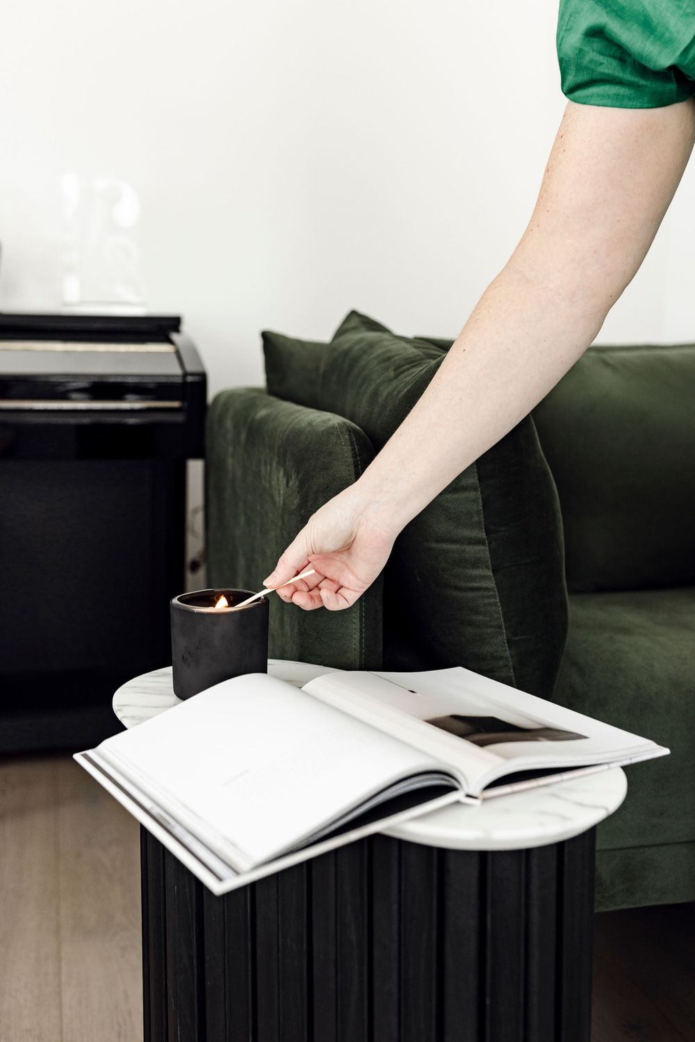 Hand lighting a candle on a marble side table beside an open magazine and green velvet sofa.